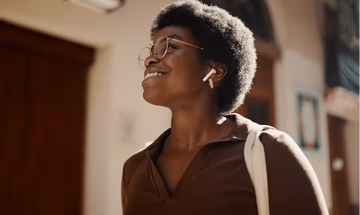Smiling woman with afro hairstyle wearing round glasses and wireless earbuds outdoors.