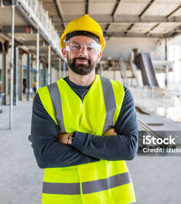 Smiling construction worker wearing a yellow hard hat, safety glasses, and a neon yellow high-visibility vest, standing with arms crossed inside a large, unfinished building site.