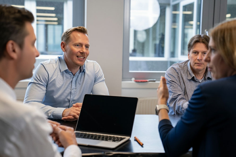 Vier Personen sitzen an einem Tisch in einem modernen Büro und führen ein Meeting, ein Laptop steht auf dem Tisch.