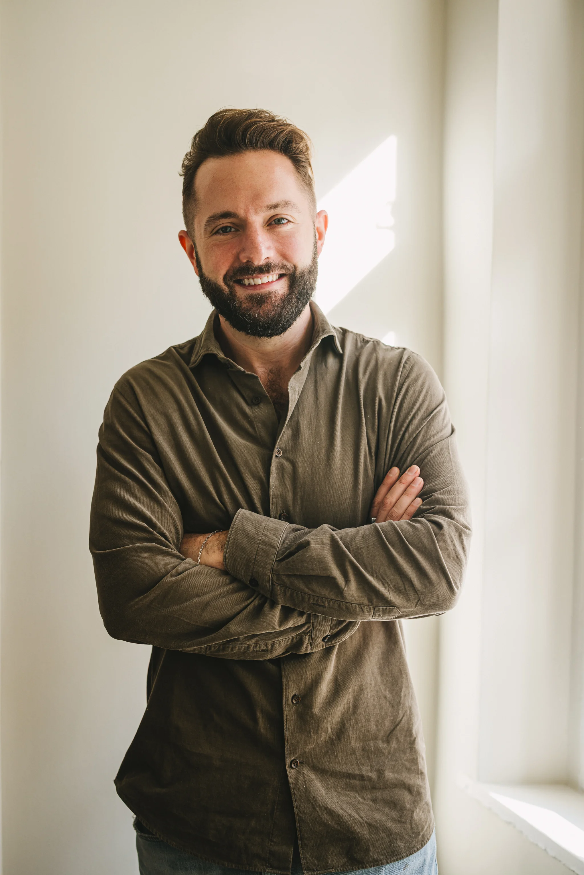 Smiling man with beard and short hair wearing a brown button-up shirt, standing with arms crossed near a sunlit window.
