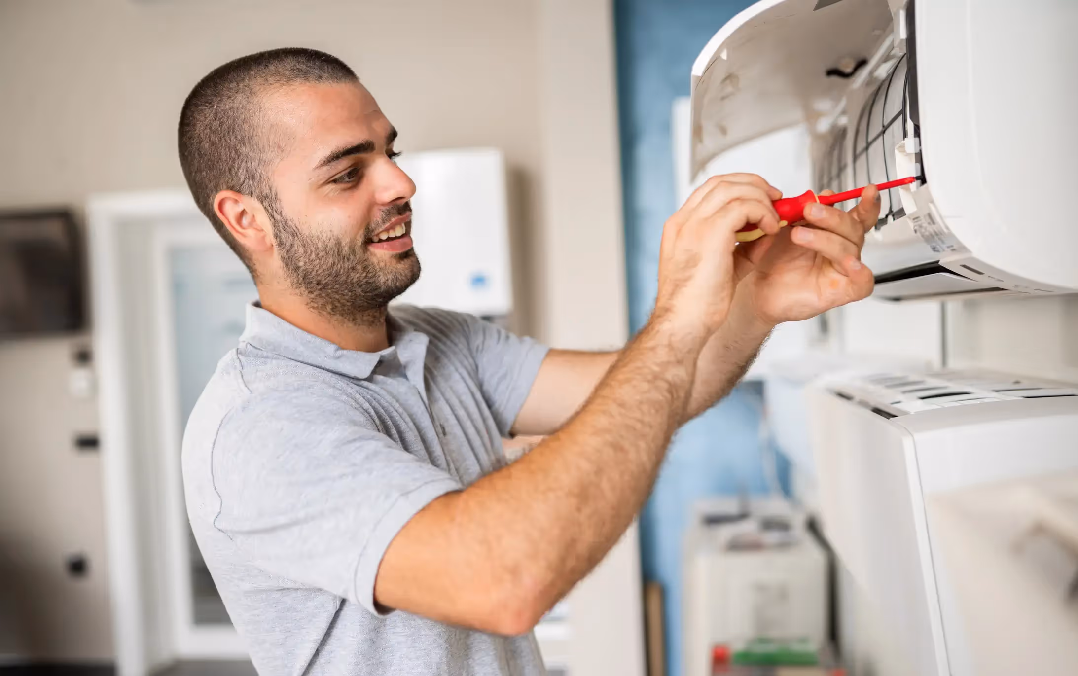 The image is a close-up shot of a smiling, young, bearded technician with a buzz cut, wearing a light grey polo shirt, as he works on an indoor, wall-mounted split-type air conditioning unit.