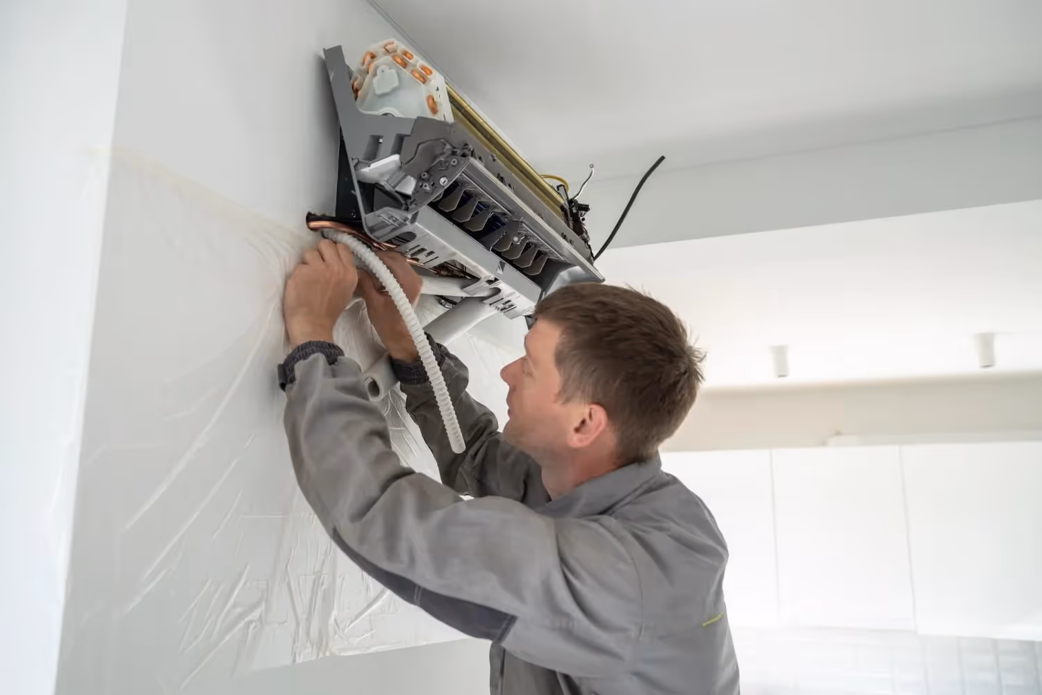An AC technician in a grey uniform working on the installation of a wall-mounted mini-split indoor unit in a bright, modern room. He is focused on connecting and routing the insulated white refrigerant lines and wiring into the back of the unit. The wall underneath the unit is partially covered by a clear plastic sheet to protect it during the installation process.