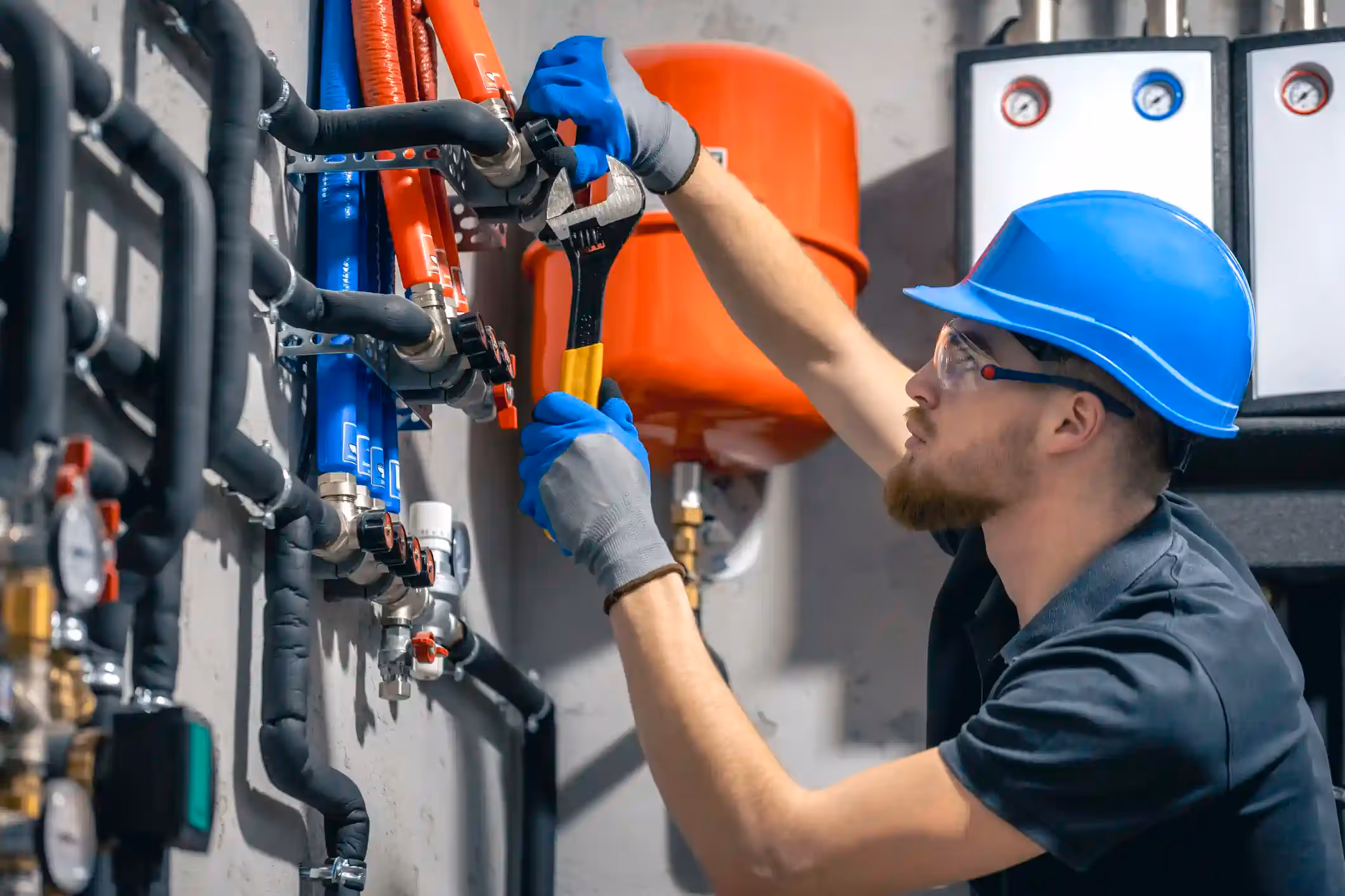 A male technician or plumber working on a complex commercial or industrial plumbing manifold system. He is wearing a dark shirt, a bright blue hard hat, safety glasses, and grey and blue gloves. He is using a wrench with a yellow handle to adjust a valve or connection on the wall-mounted piping, which includes insulated black pipes, and red and blue plastic pipes (likely for hot and cold water or radiant heating). A large red expansion tank is visible in the background, along with white panels featuring pressure gauges.