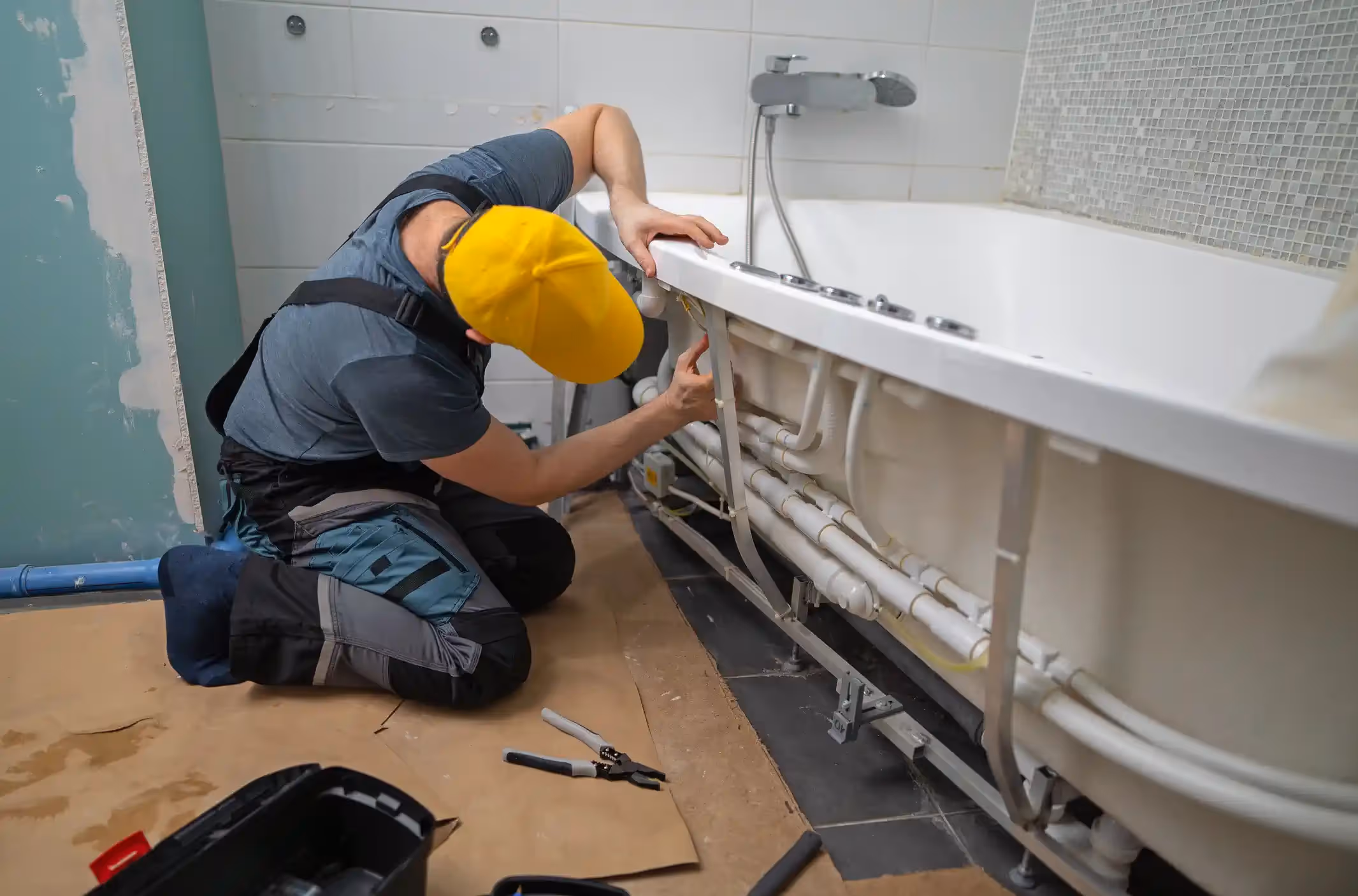 A plumber or installer kneeling on the floor, which is partially covered with brown protective paper, while working on the plumbing connections for a bathtub. He is wearing a dark grey shirt, two-toned work pants (grey and black), a yellow baseball cap, and is focused on connecting or adjusting the numerous white plastic pipes and wiring visible underneath the tub. The bathroom has white tiled walls, and the tub itself is white with a mounted faucet.
