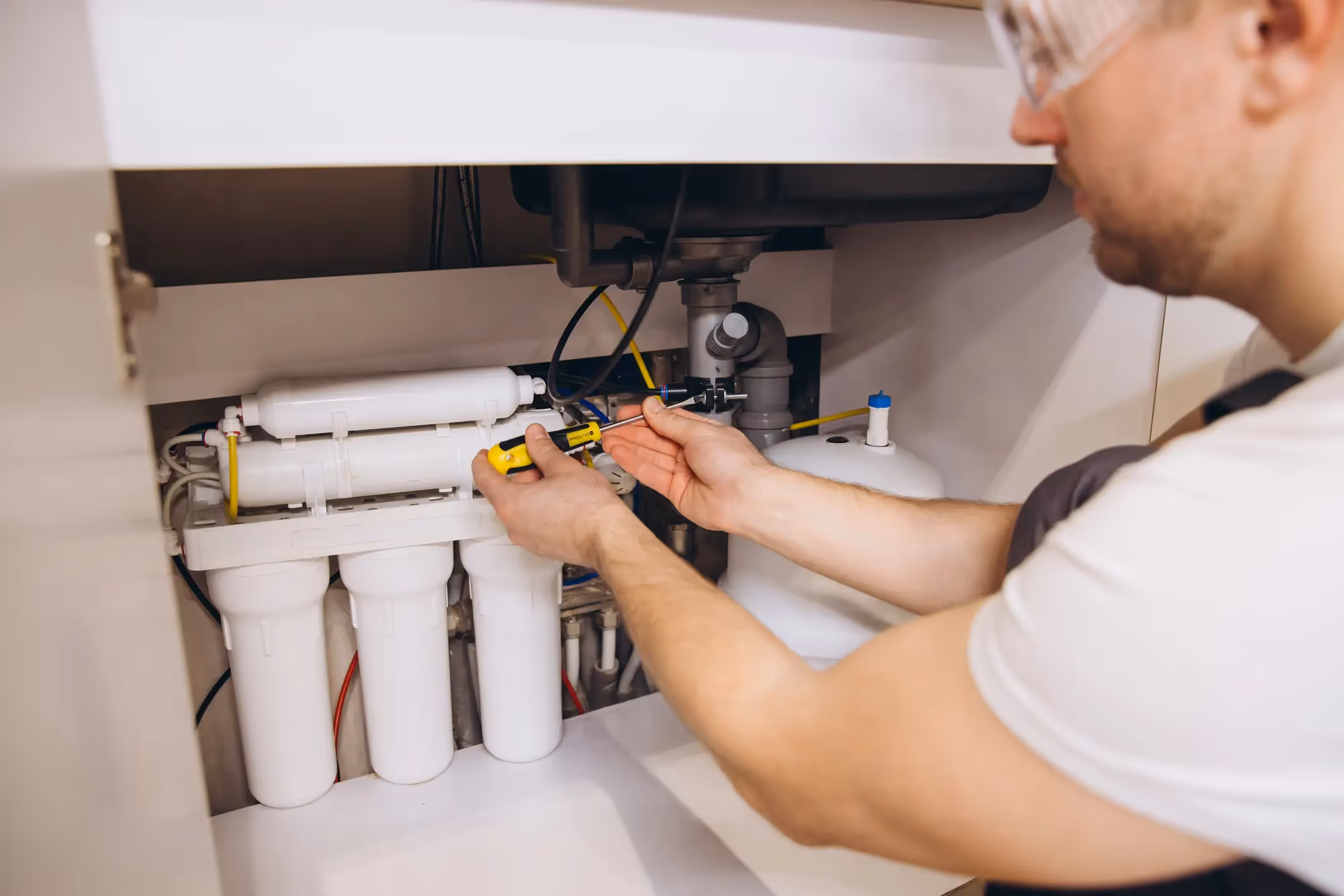 A technician wearing a white shirt and clear safety glasses, installing or servicing an under-sink Reverse Osmosis (RO) water filtration system. He is leaning into the open cabinet below the sink and using a small yellow and black screwdriver to work on the tubing or connections of the white filtration unit. The system has four visible filter housings and a white pressure tank to the right.
