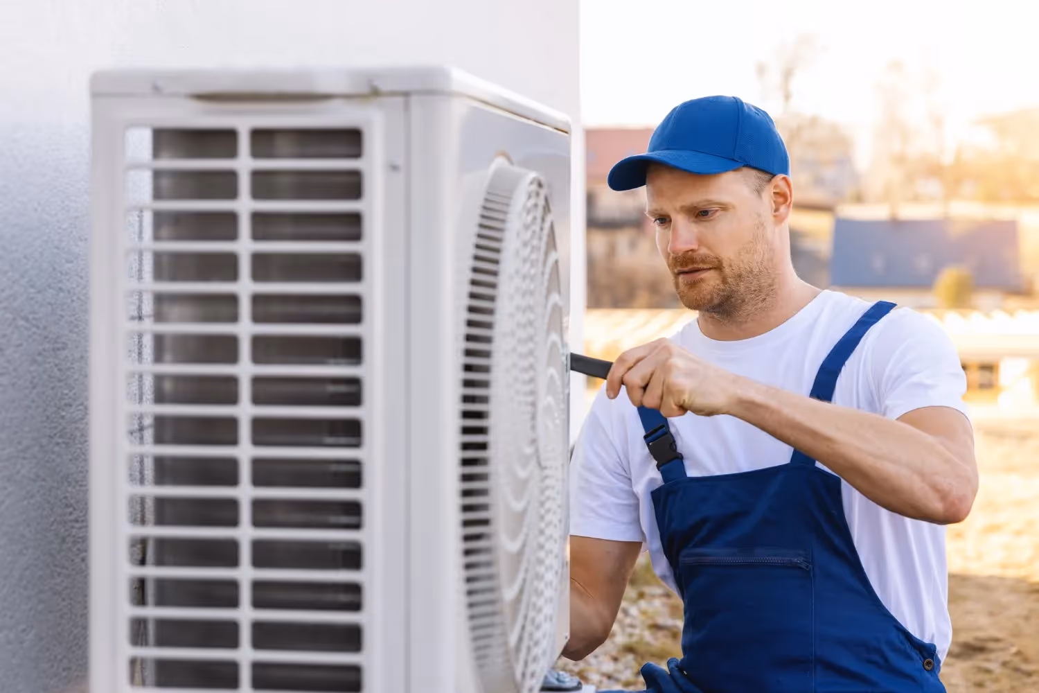 A man wearing a white t-shirt, blue overalls, and a blue baseball cap is focused on working on an outdoor air conditioning or heat pump unit. He appears to be tightening or adjusting a component on the side of the white unit using a dark tool, possibly a wrench or screwdriver. The background is a blurred suburban setting with sunny lighting.