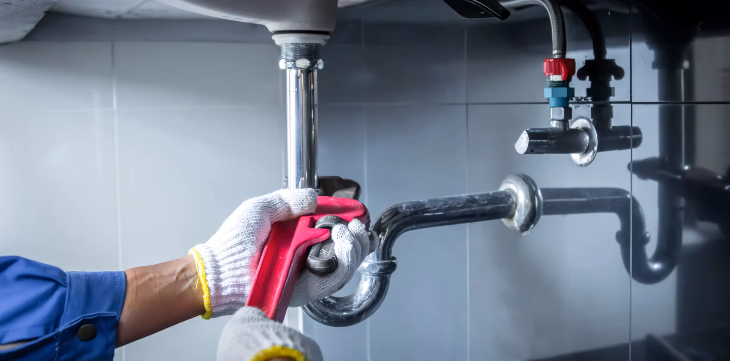 A close-up image shows a plumber, wearing white and yellow work gloves and a blue uniform sleeve, using a red pipe wrench to adjust or tighten a chrome P-trap fitting beneath a sink. The drainpipe curves upward from the trap and connects to the sink's basin. In the background, water supply lines with red (hot) and blue (cold) shut-off valves are visible against a dark, tiled wall.