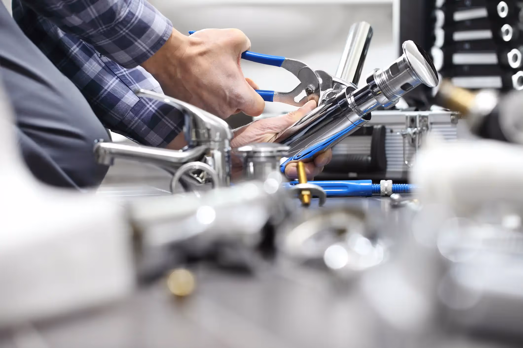 A close-up, low-angle shot shows a plumber's hands, wearing a plaid shirt, using blue-handled adjustable pliers to tighten or assemble a shiny chrome faucet component on a dark surface. Several other chrome plumbing parts, including a completed sink faucet, are scattered and blurred in the foreground and background, suggesting a workspace for commercial fixture installation or repair.