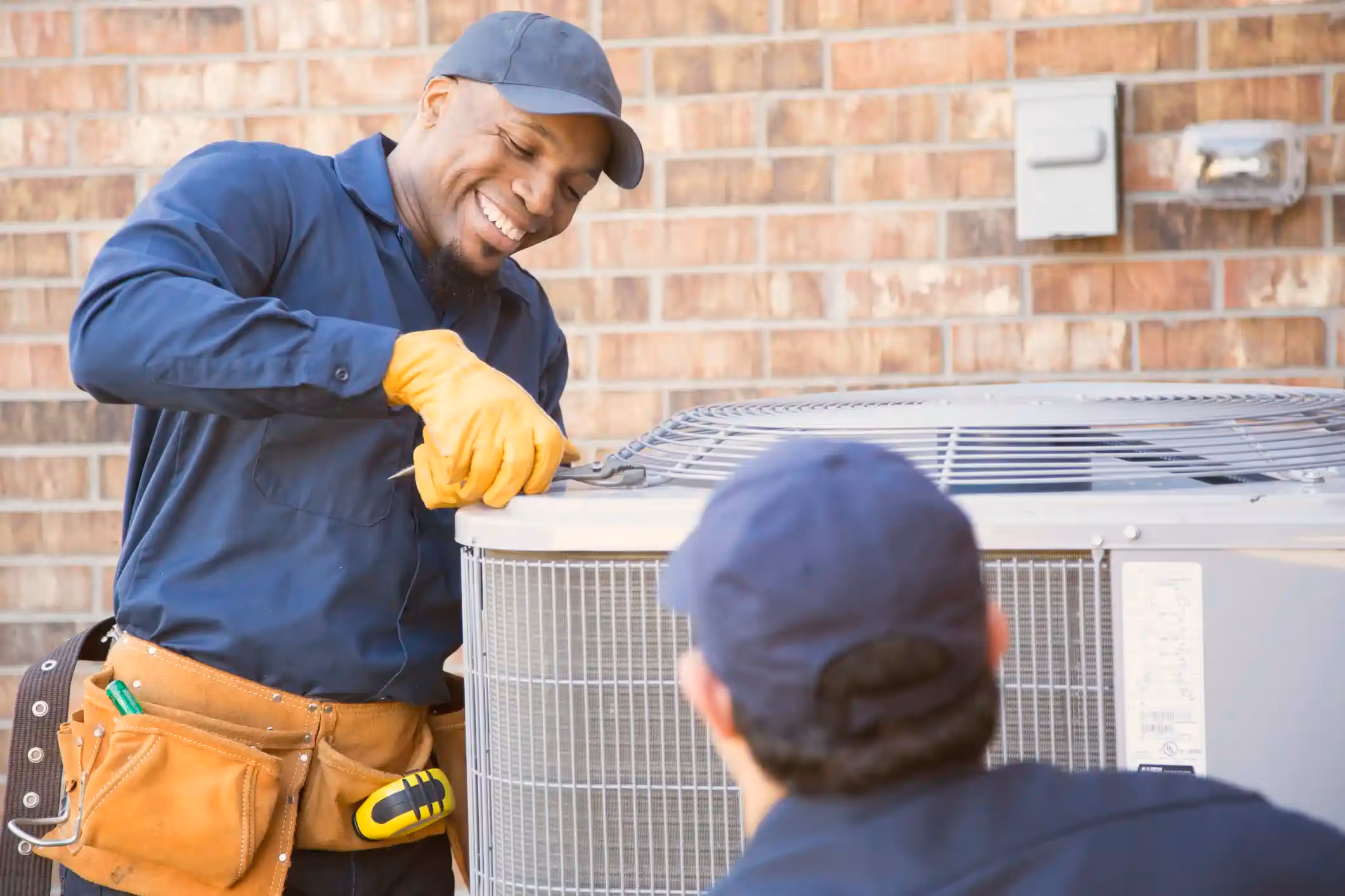 A cropped, close-up image shows a smiling Black HVAC technician in a dark blue uniform, cap, and a leather tool belt, working on the top of an outdoor air conditioning unit. He is wearing bright yellow gloves and using a wrench. Another technician, whose back and blue cap are visible in the foreground, is also working on the unit against a red brick wall.