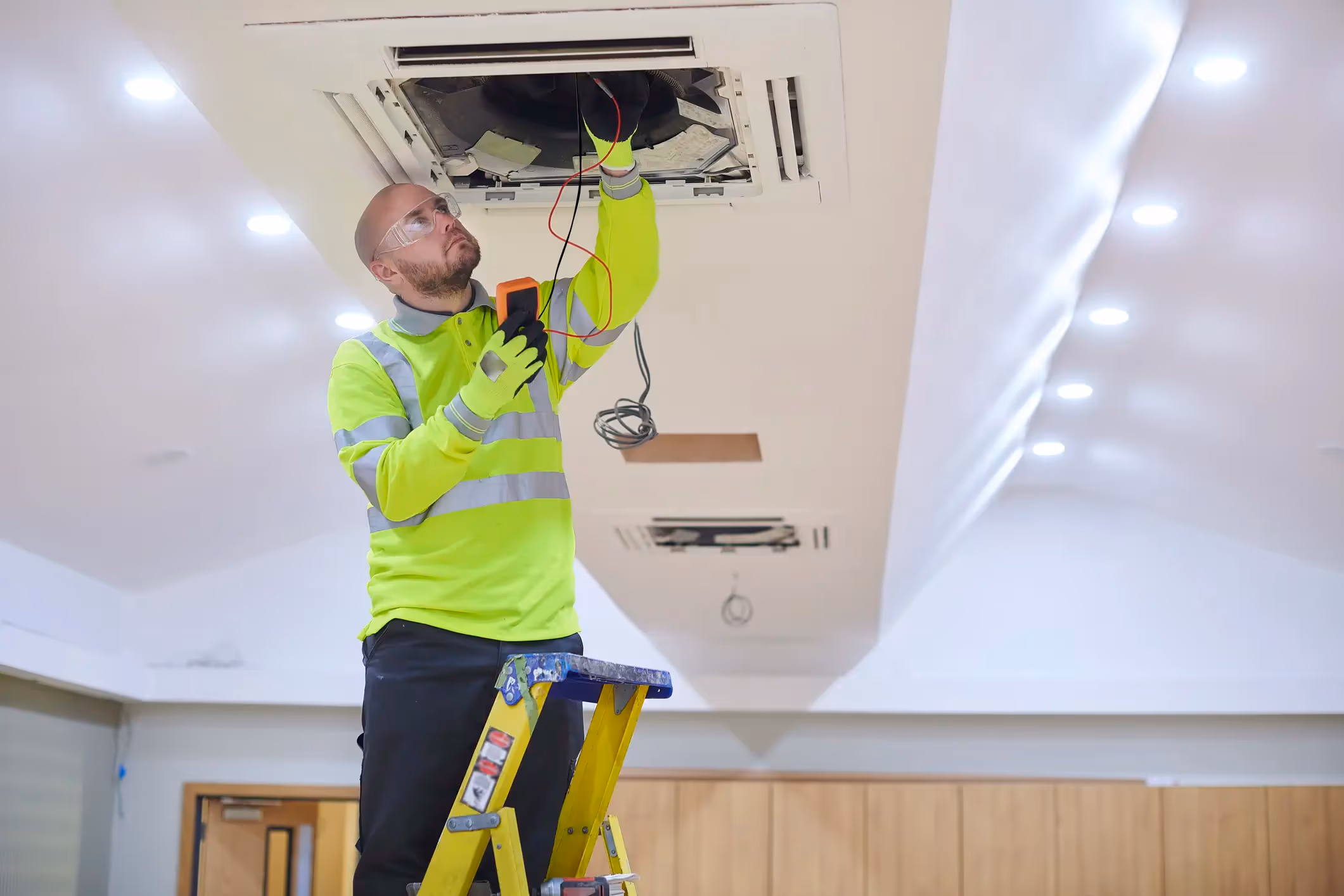 A man, wearing a high-visibility yellow-green shirt, black pants, and safety glasses, is standing on a yellow ladder and servicing a ceiling-mounted cassette air conditioning unit. He is using a multimeter to check the wiring or electrical components of the open unit. The room has a white ceiling with multiple recessed lights and wooden paneling in the background.