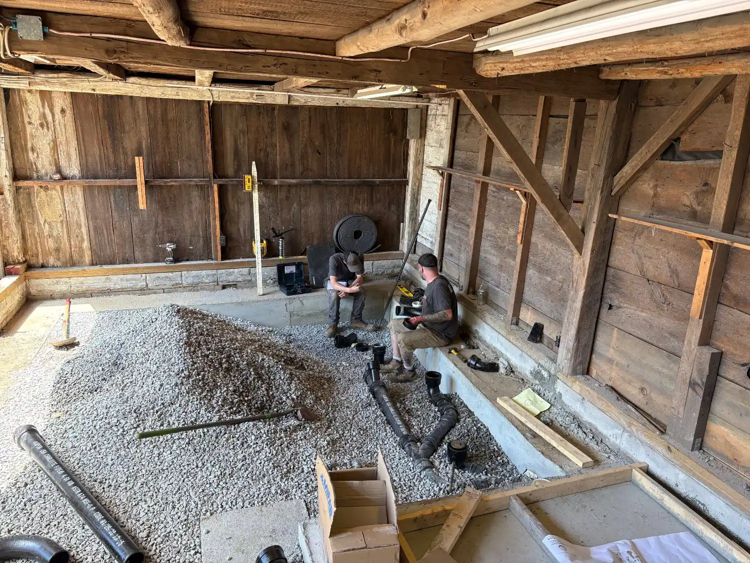 Two plumbers sit on a low concrete ledge inside a rustic, timber-framed barn undergoing renovation. They are assembling black PVC drainage pipes on a floor covered in gray gravel, surrounded by construction tools and materials.
