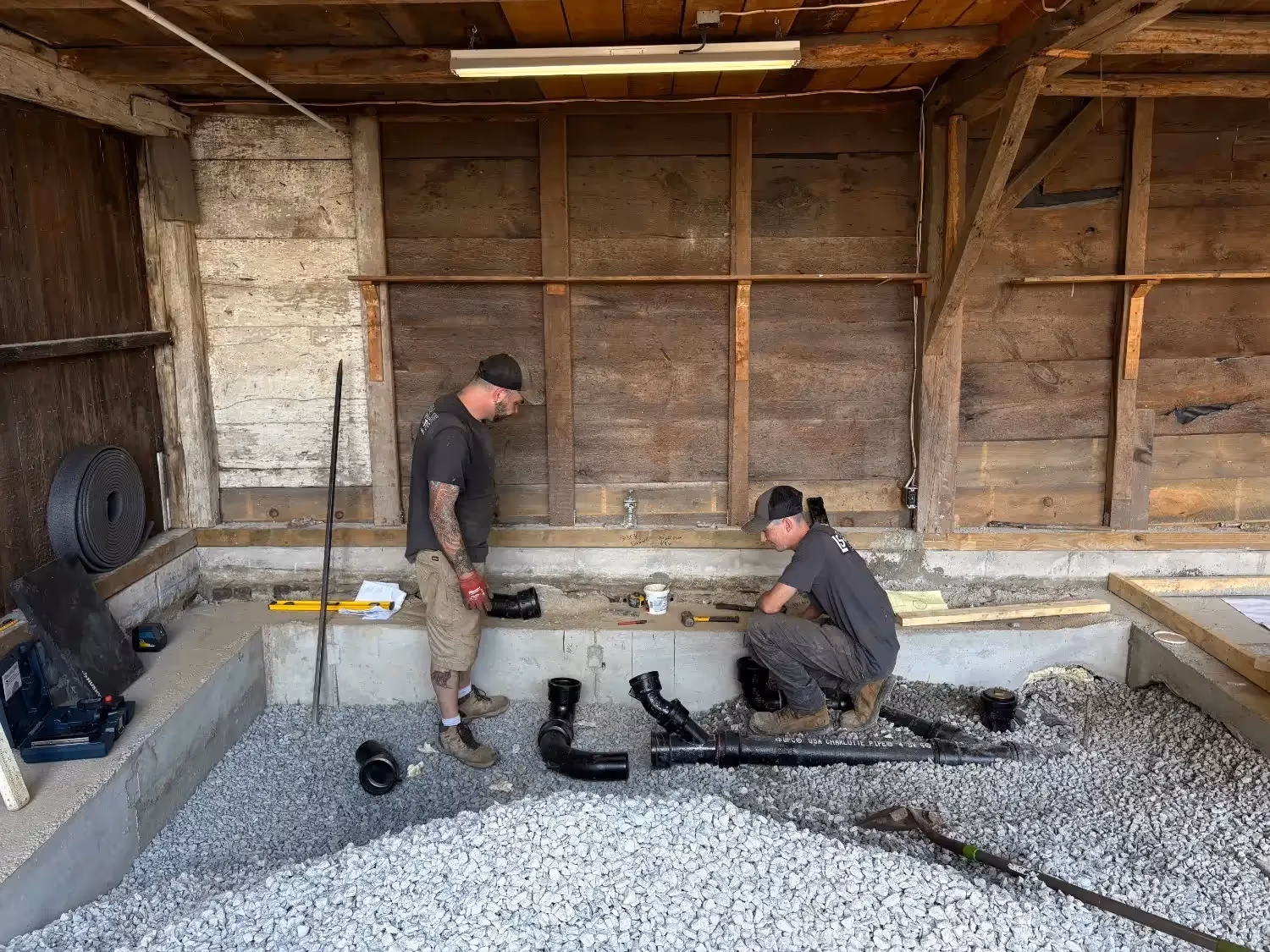 Two plumbers in a rustic, wood-walled barn work on black cast iron or PVC drainage pipes set into a bed of gray gravel. One worker stands while the other crouches near a concrete foundation ledge, surrounded by construction tools and materials.