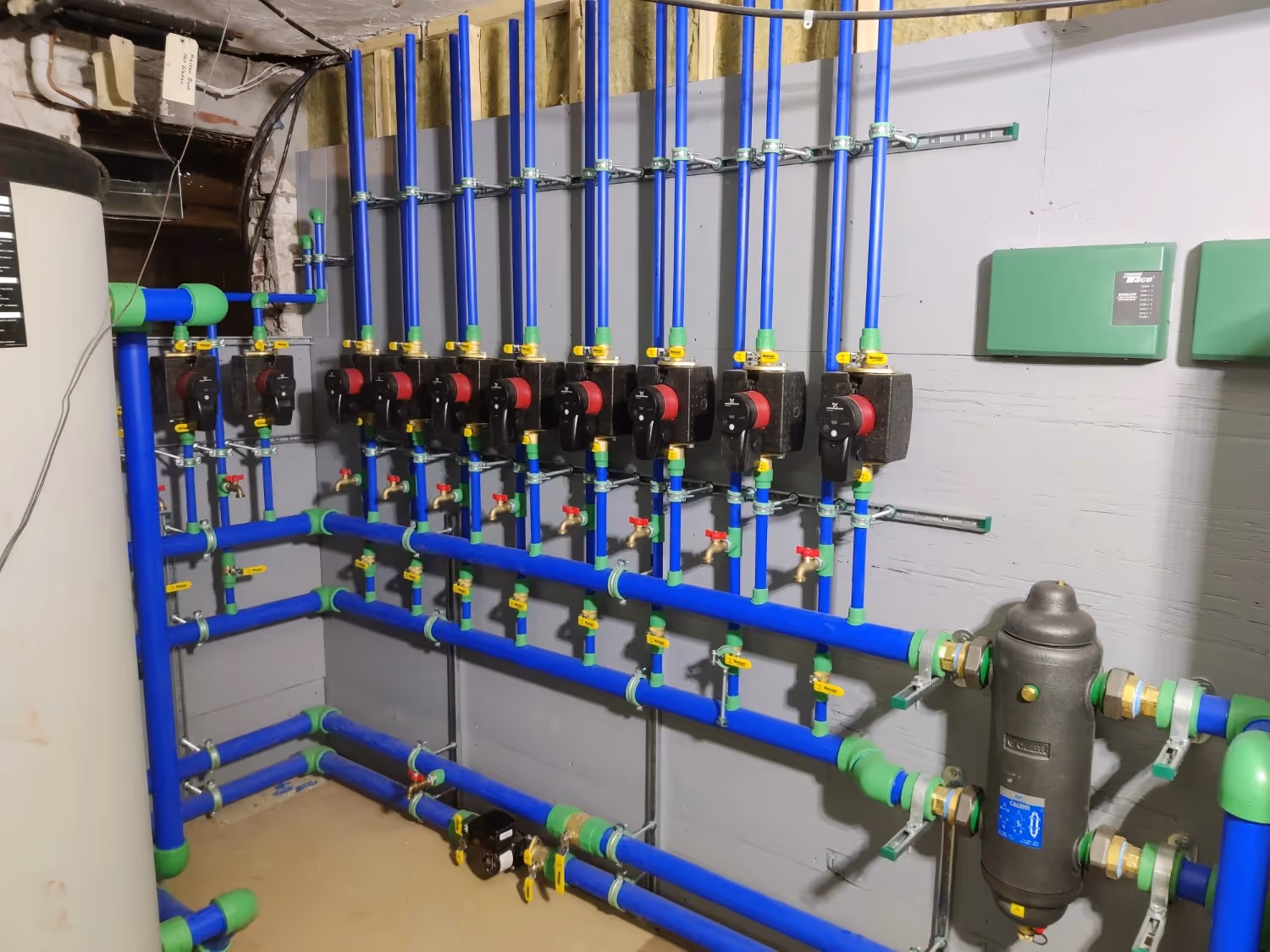 A complex hydronic manifold system featuring vibrant blue and green Aquatherm piping mounted to a wall. Multiple black Grundfos circulation pumps are arranged in a row with red accents, accompanied by various brass valves, gauges