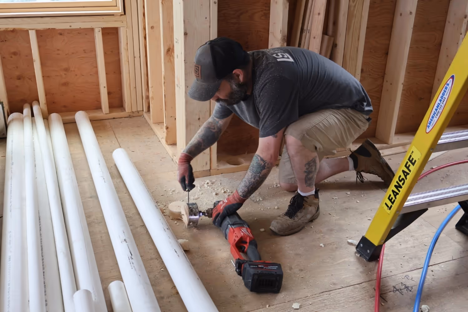 A worker with tattoos kneels on a plywood floor at a construction site, using a red Milwaukee right-angle drill to cut a hole. Several white PVC pipes lie on the floor nearby, and a yellow ladder is partially visible on the right.