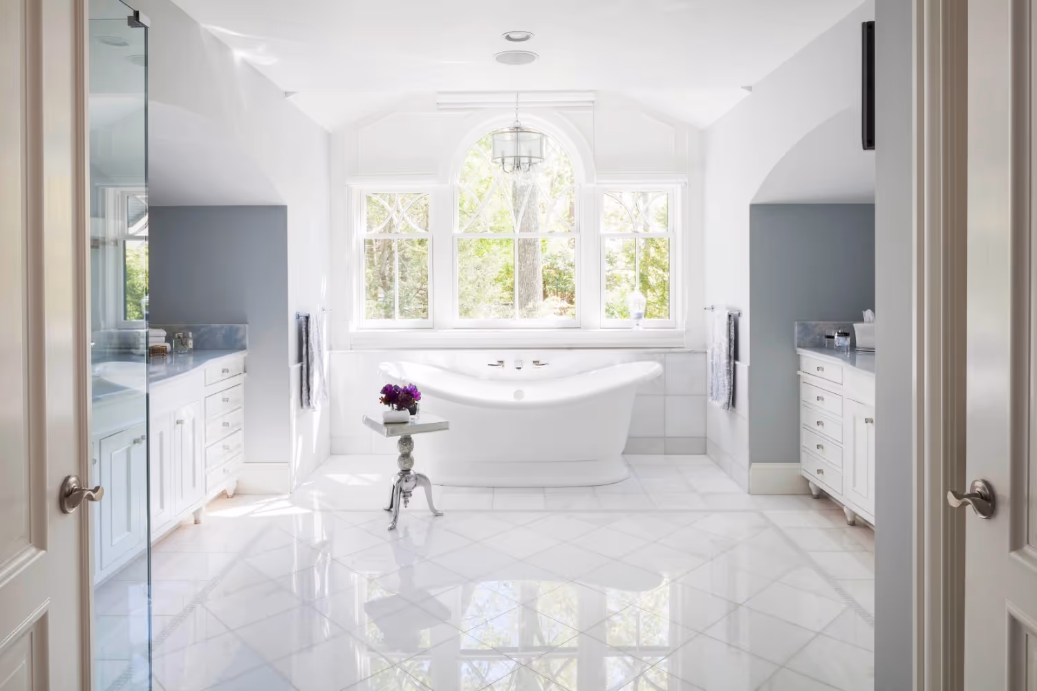 A spacious, luxurious bathroom featuring polished white marble floors with a diamond pattern and a central freestanding soaking tub positioned beneath a large arched window.