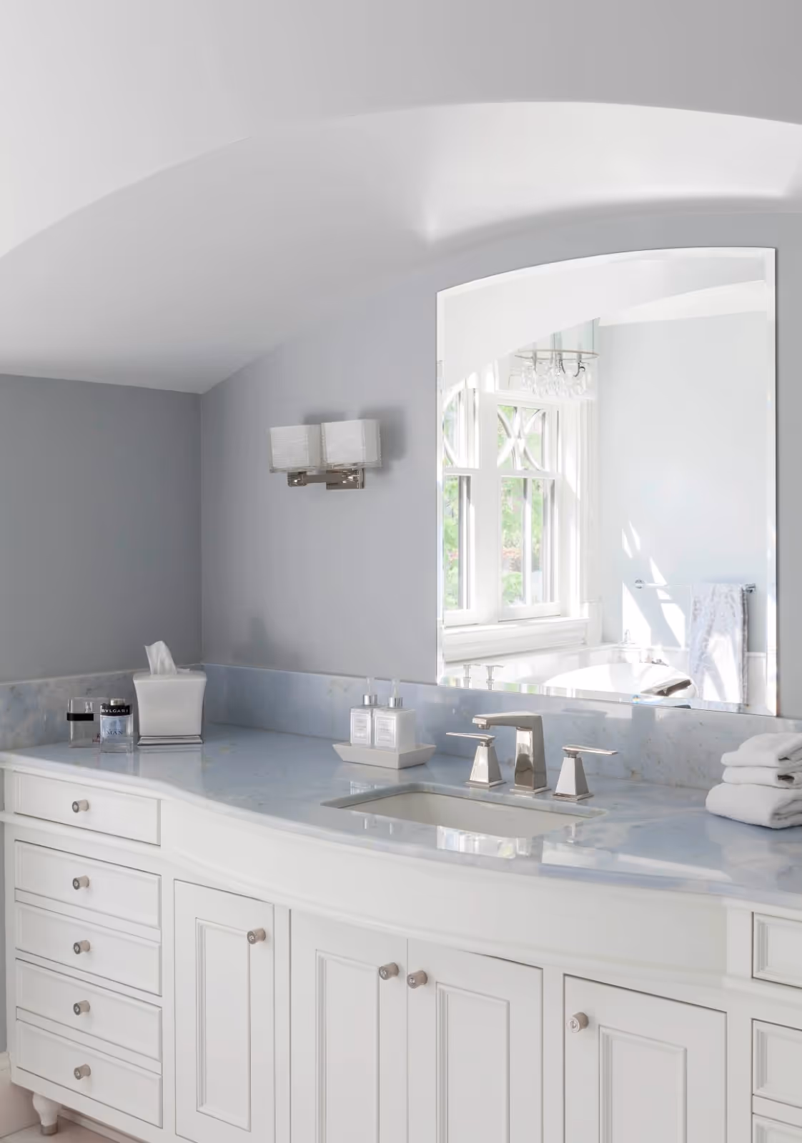 A white shaker-style bathroom vanity features a light blue marble countertop and chrome faucet. An arched frameless mirror is mounted on a grey wall beneath a sloped white ceiling, reflecting a bright window and chandelier.