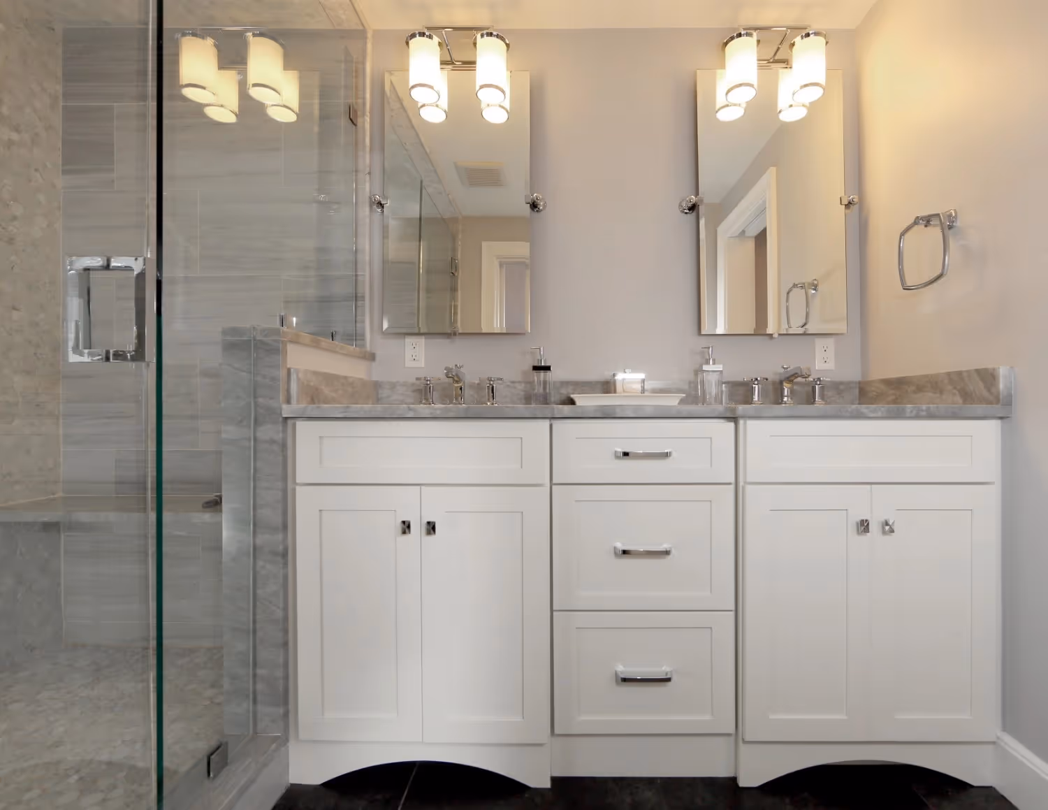 A double vanity in a contemporary bathroom features white shaker-style cabinets with a grey marble countertop and two undermount sinks. Above the vanity are two frameless mirrors flanked by modern chrome light fixtures. A glass-enclosed shower with grey tile and a stone bench is visible to the left.