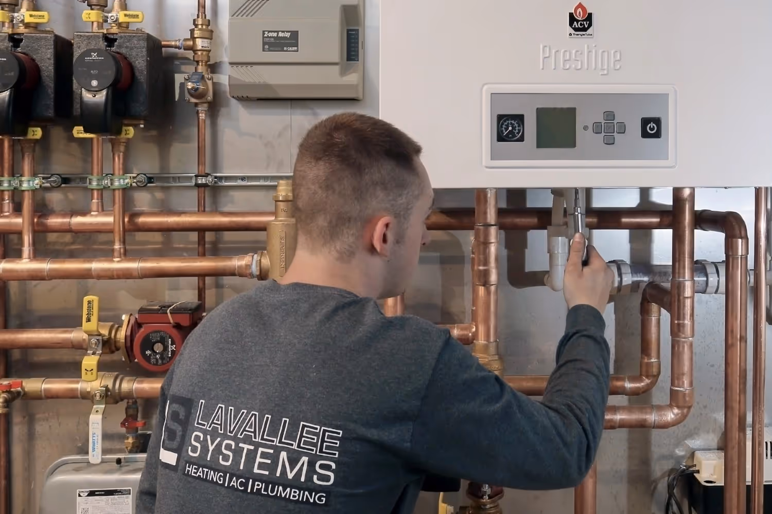 A technician wearing a Lavallee Systems shirt performs maintenance on a white Triangle Tube Prestige boiler. The unit is connected to an intricate copper piping manifold featuring several black and red Grundfos circulation pumps, a Caleffi Z-one relay, and various brass fittings and valves.
