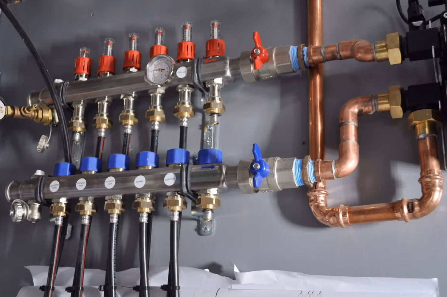 A radiant heating manifold is wall-mounted and connected to copper piping. It features two stainless steel headers with red flow meters on top and blue actuators on the bottom. Several black PEX tubes extend downward, each labeled for specific zones like "recreation room" and "bathroom."