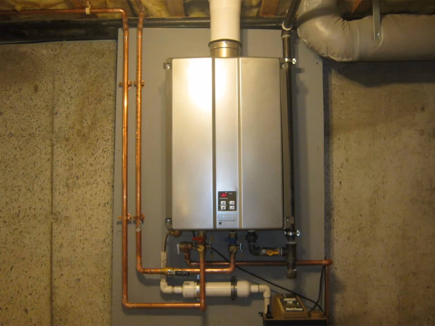 A sleek, wall-mounted Rinnai tankless water heater is installed on a grey panel in a basement. The unit features a digital temperature display set to 120°F and is connected to a network of copper piping, a white PVC vent, and a condensate pump.