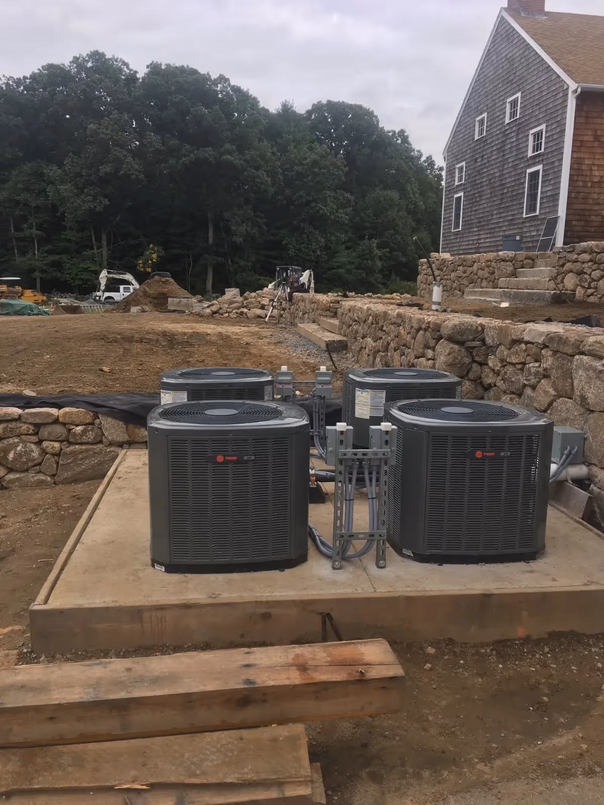 Four grey Trane central air conditioning condenser units are installed on a concrete pad at a construction site. The outdoor area features stone retaining walls, a large shingled house in the background, and excavation equipment visible near a wooded area.