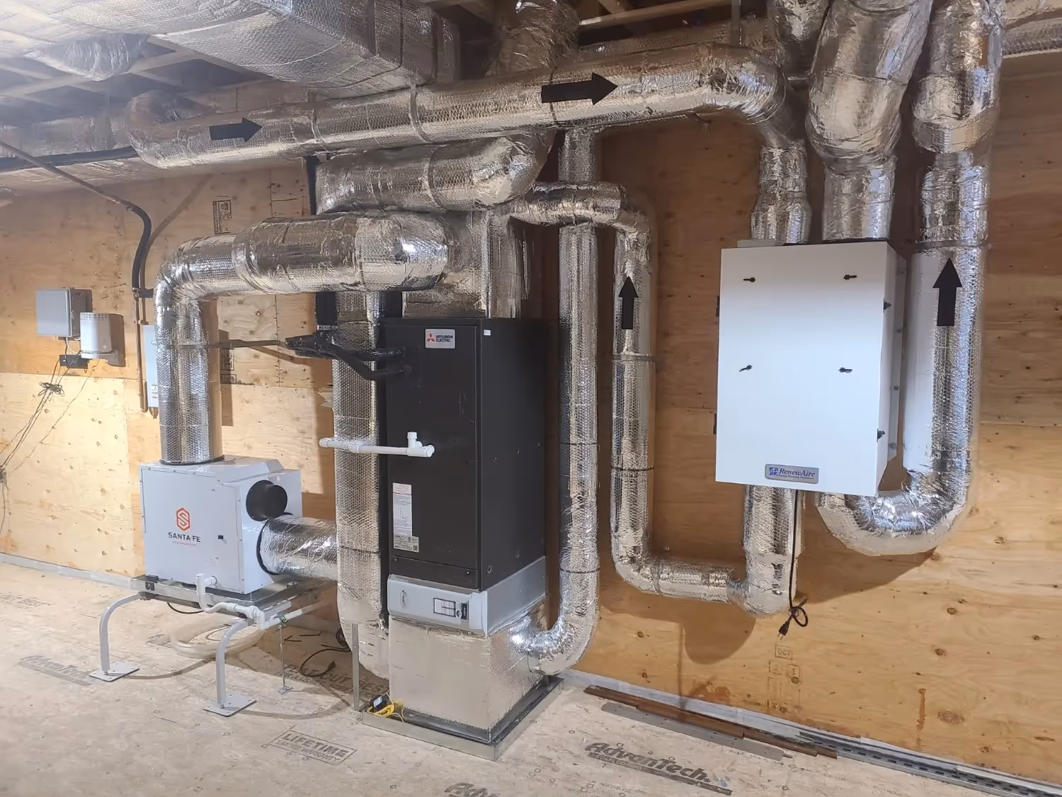 Two white Mitsubishi Electric outdoor condenser units are installed on metal stands against a red brick and grey foundation wall. A grey conduit runs between the units, which are surrounded by grey gravel and green foliage.