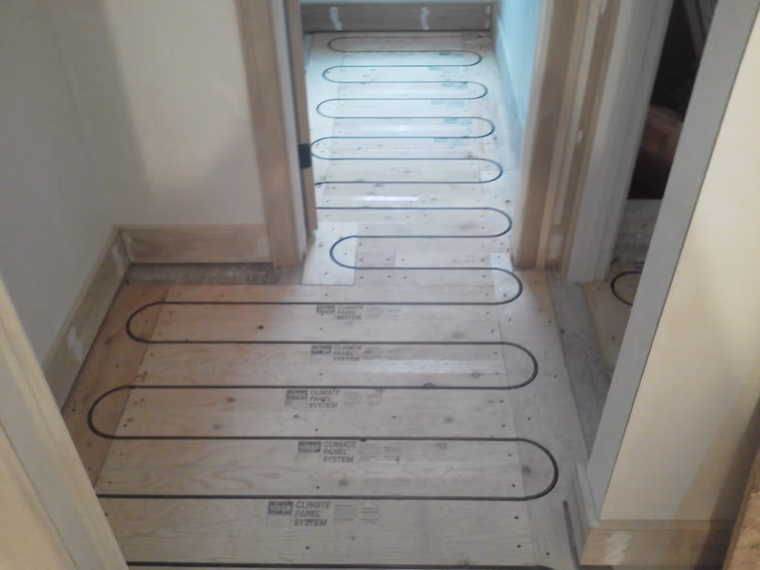 A network of black radiant heating tubes is installed in a serpentine pattern across a plywood subfloor in a hallway and adjacent rooms. The wooden "Climate Panel System" boards feature pre-cut grooves to hold the tubing securely before the final flooring is installed.
