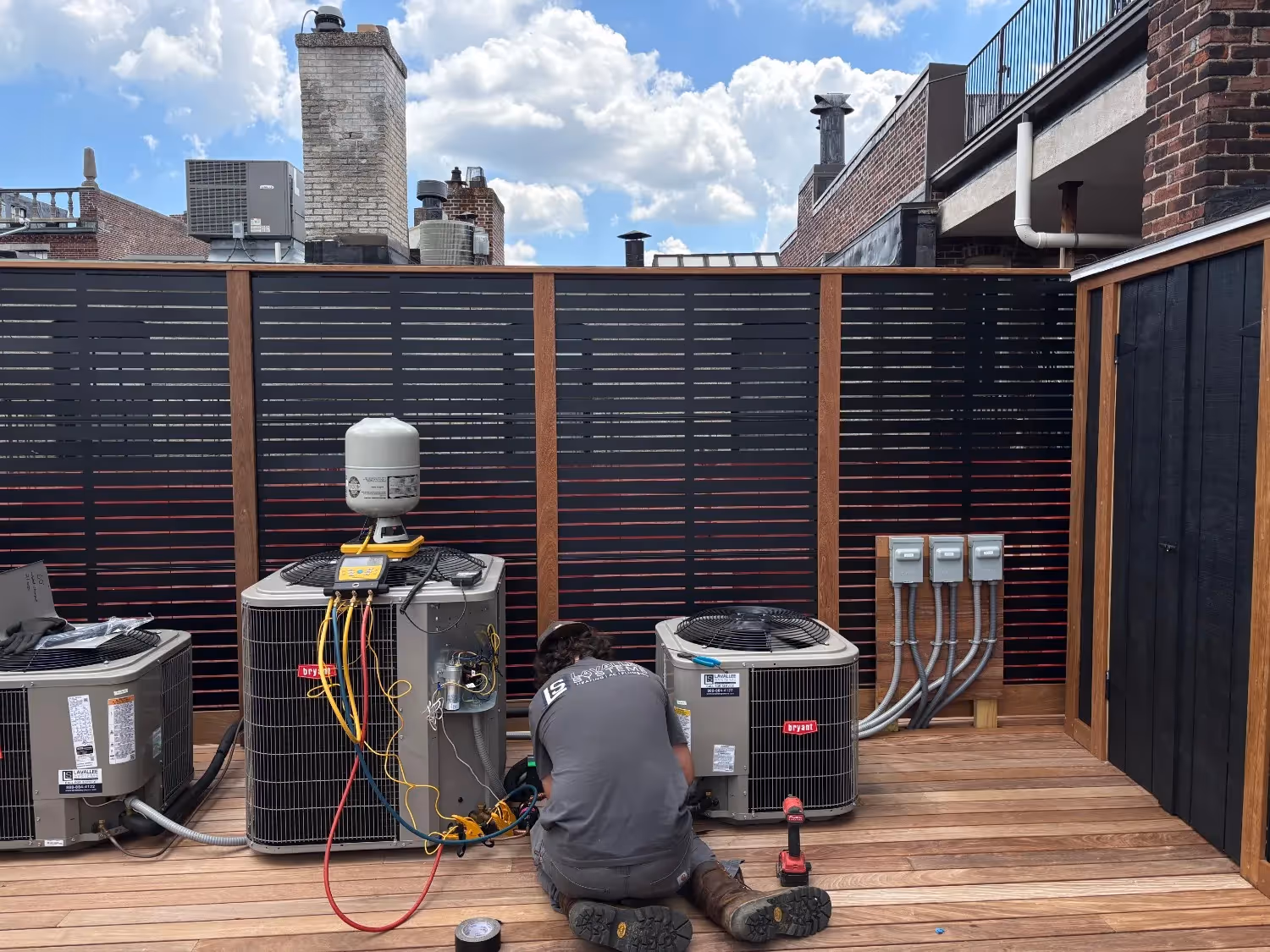 A technician sits on a wooden rooftop deck, performing maintenance on three Bryant AC units. The technician wears a grey Lavallee Systems shirt and uses a digital manifold gauge with colorful hoses to test the central unit. A black slatted privacy fence stands in the background.