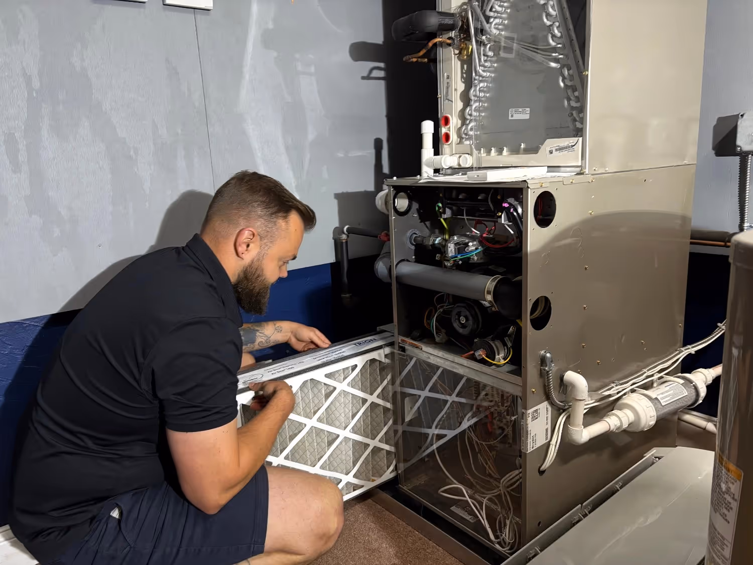 A technician with a beard and arm tattoos kneels on the floor, sliding a new white pleated air filter into the base of an open HVAC furnace unit. The unit’s internal components, including wiring and coils, are visible as he performs the maintenance.