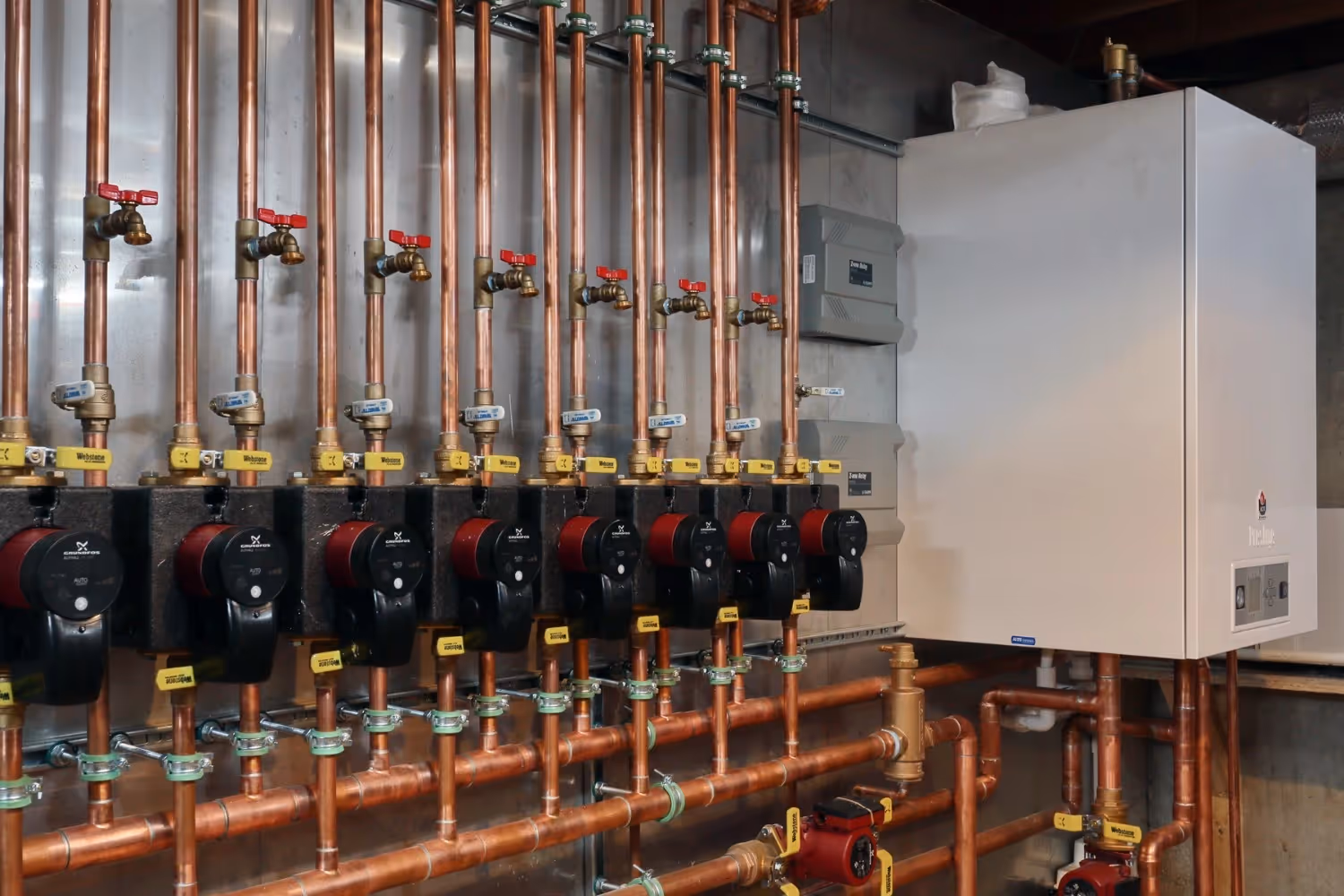 A professional mechanical setup features a wall-mounted Triangle Tube boiler connected to an extensive copper piping manifold. Eight black and red Grundfos circulation pumps are arranged in a row, each equipped with yellow-handled shut-off valves and brass drain faucets against a sleek metallic wall panel.