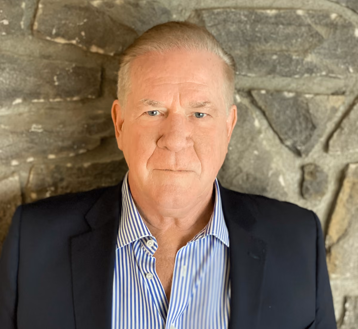 Portrait of an older man with short hair wearing a black blazer and striped shirt against a stone wall.