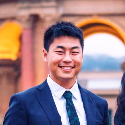 Young man in a dark suit and patterned tie smiling outdoors with an arched structure in the background.
