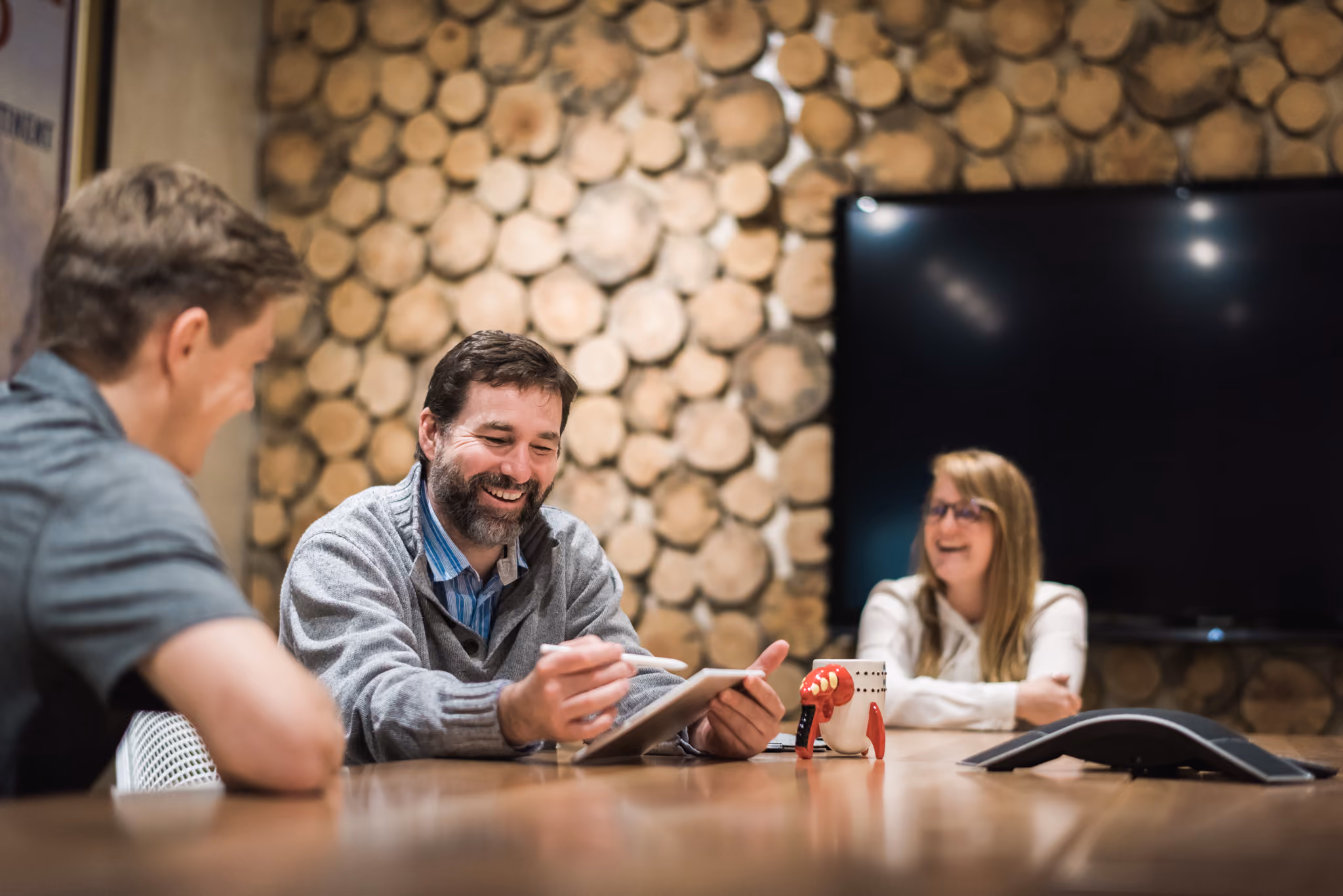Three colleagues smiling and talking around a board room table with a tablet and mug present.