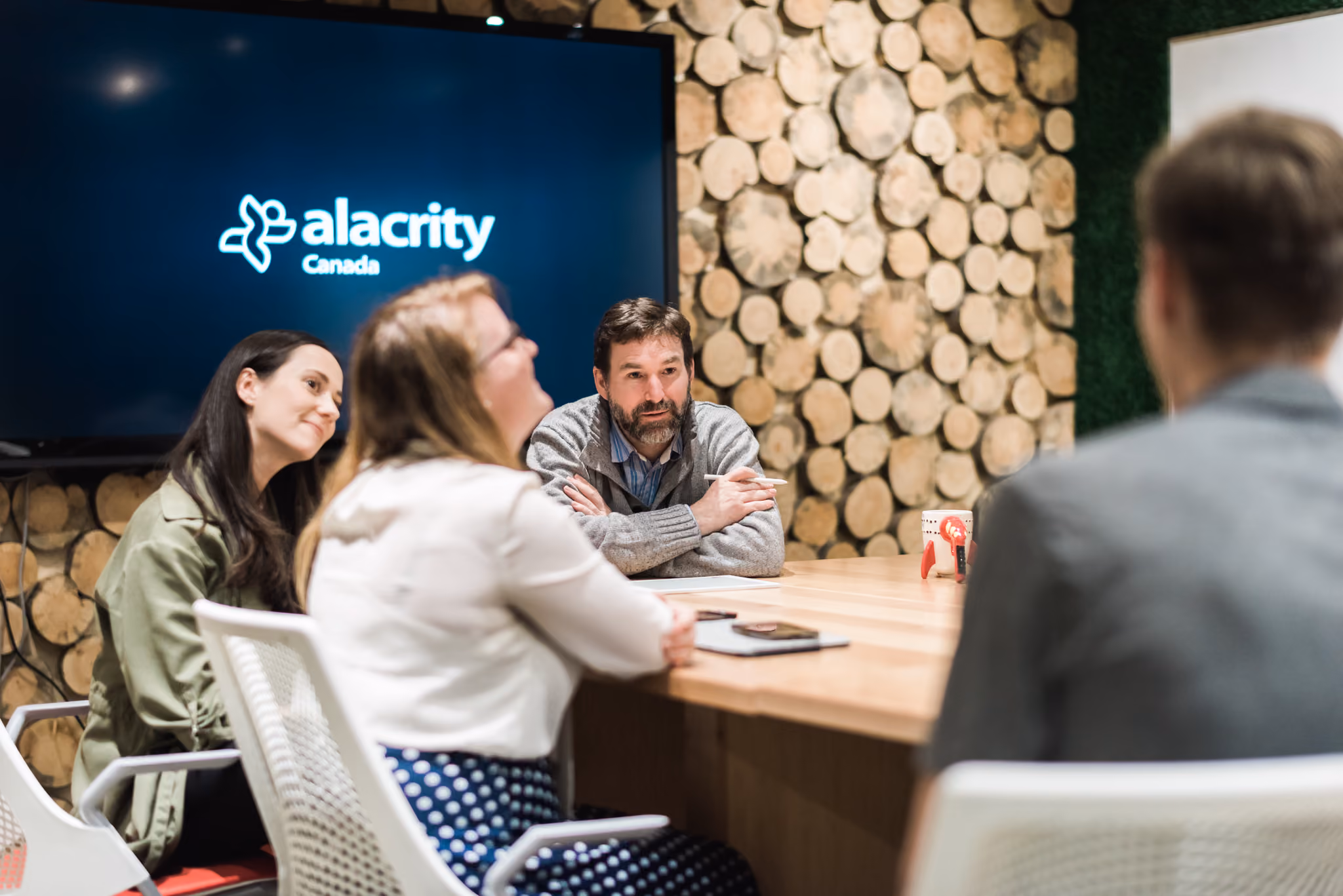 Four people having a meeting around a wooden table with Alacrity Canada logo on screen.