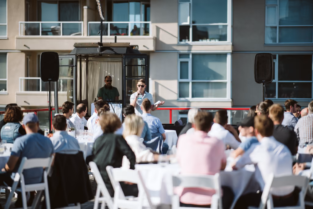 Man speaking with microphone to seated audience at outdoor event in front of apartment balconies.