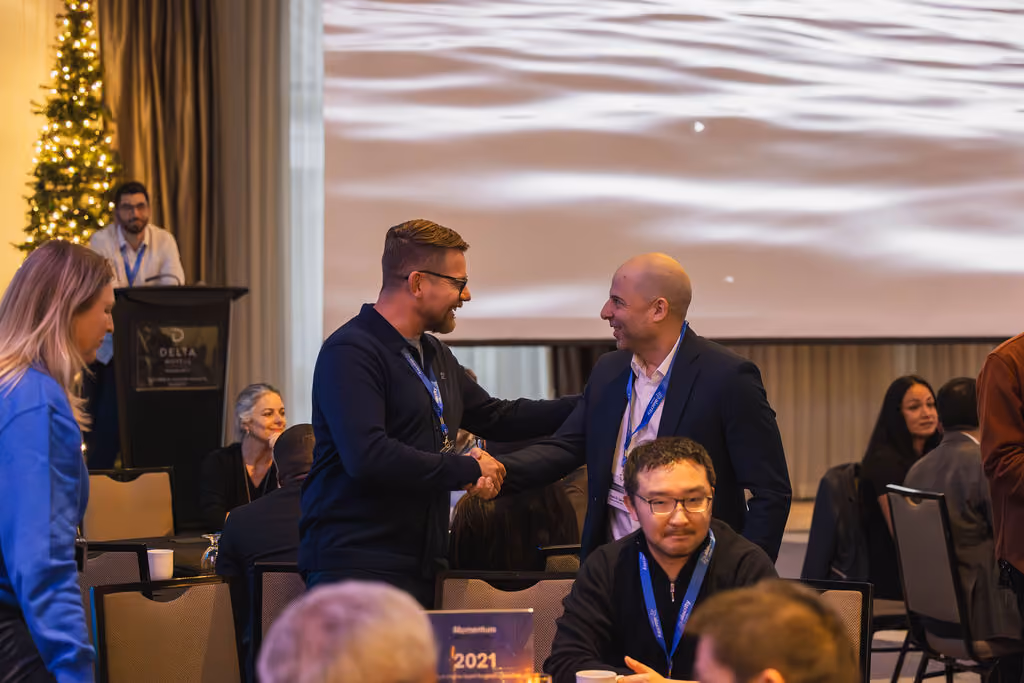 Two men shaking hands and smiling at a conference event with guests seated around them.