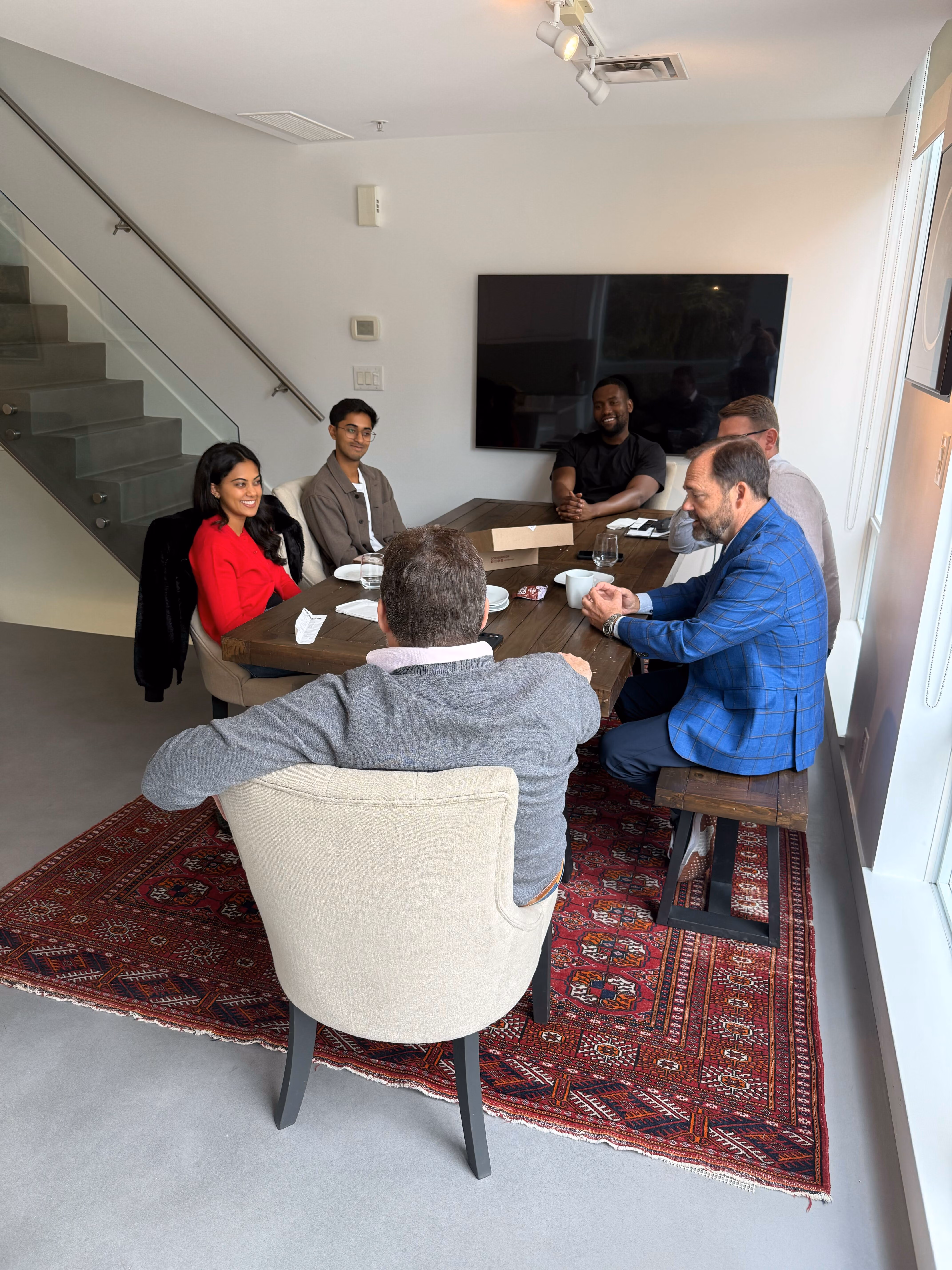 Six people sit around a wooden table in a modern room with a red rug and stairs nearby.