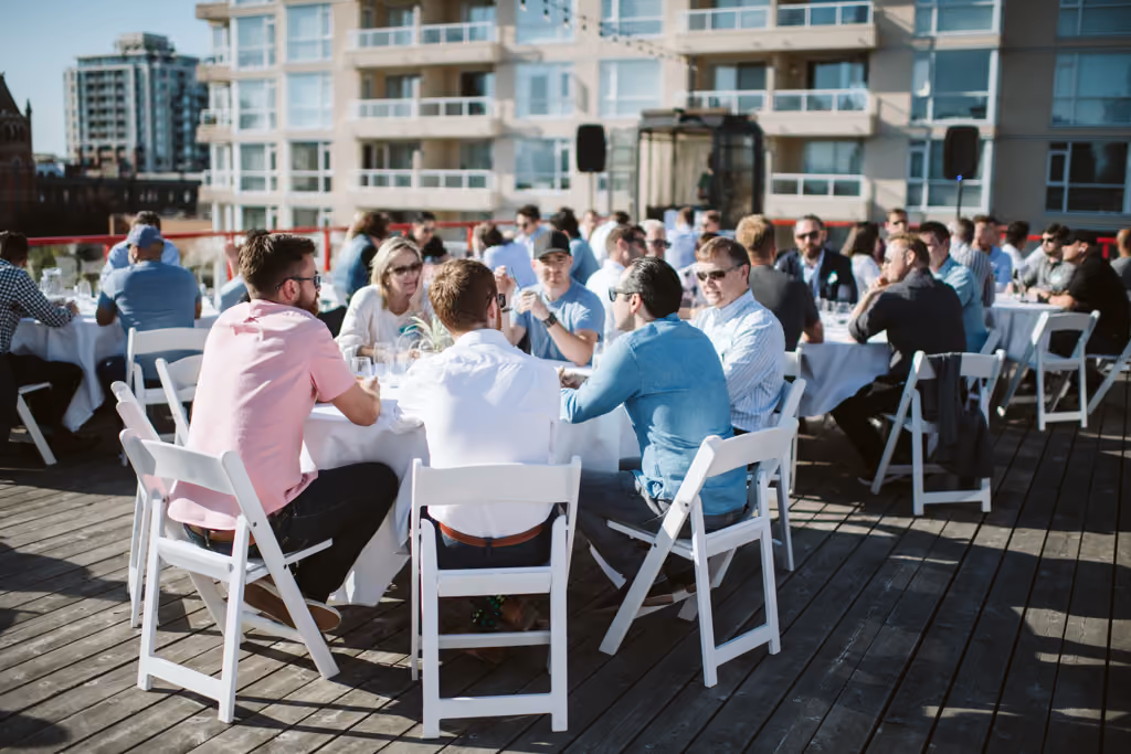 People seated at round tables outdoors on a wooden deck with modern buildings in the background.