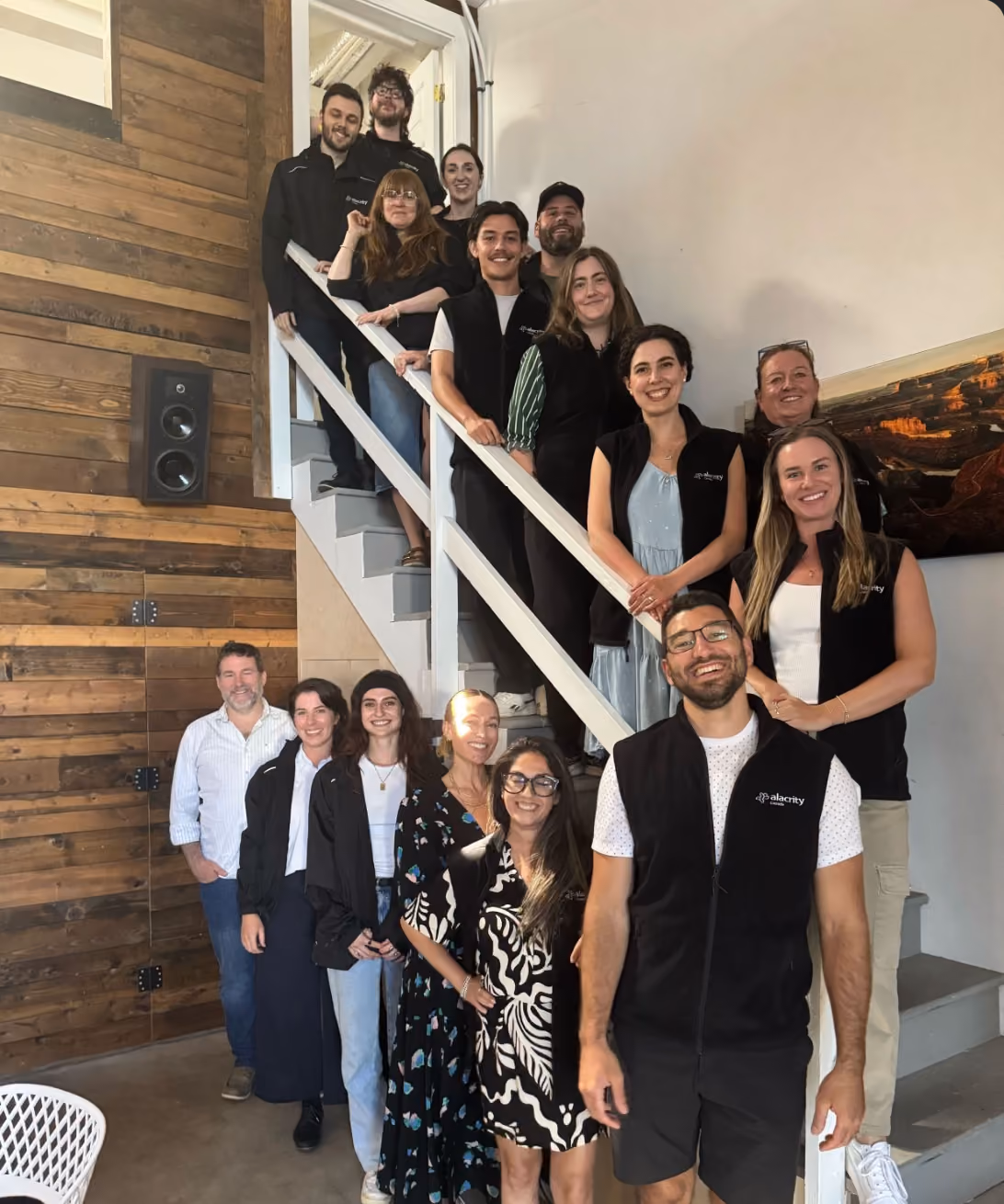Group of fifteen people smiling on and around a staircase indoors next to a wooden wall with a speaker.