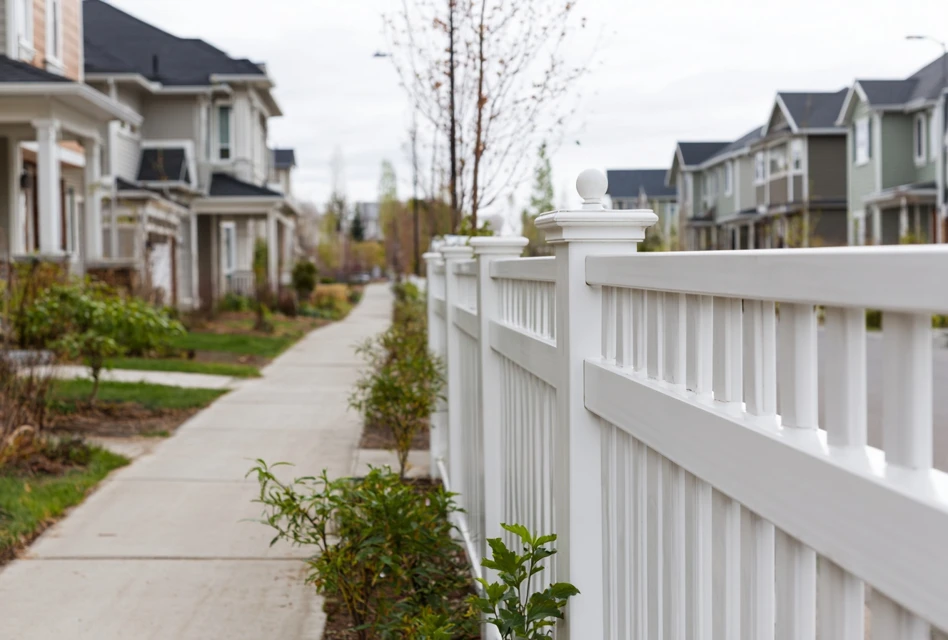 White wooden fence lining a sidewalk in a suburban neighborhood with houses and small trees.