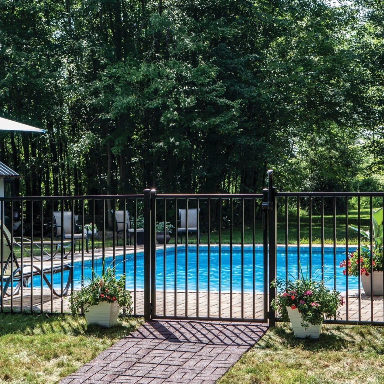 Black metal fence gate enclosing a backyard swimming pool with two flower pots on either side and lawn chairs in the background.