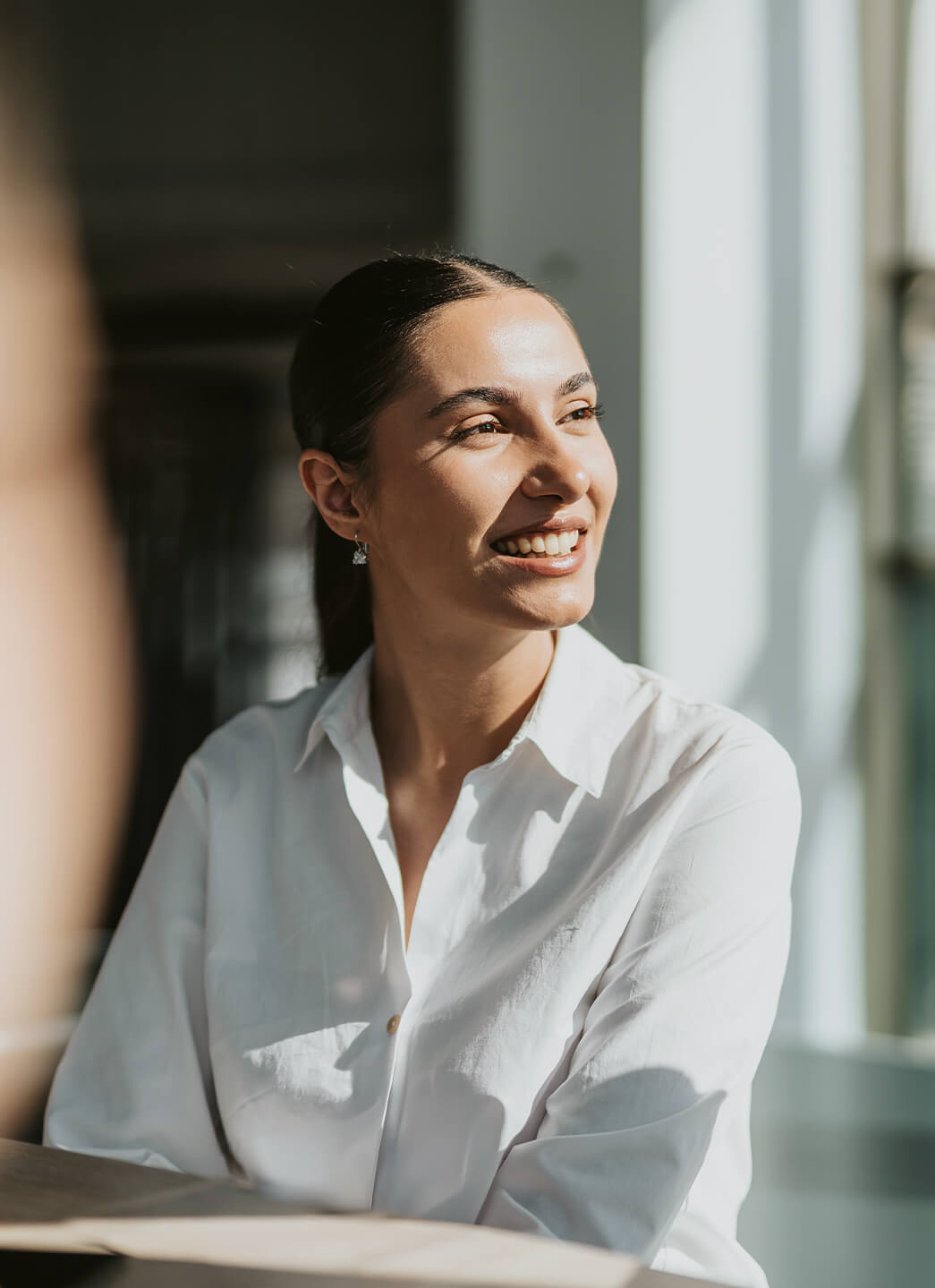 Smiling woman in a white shirt looking out a window with sunlight on her face.