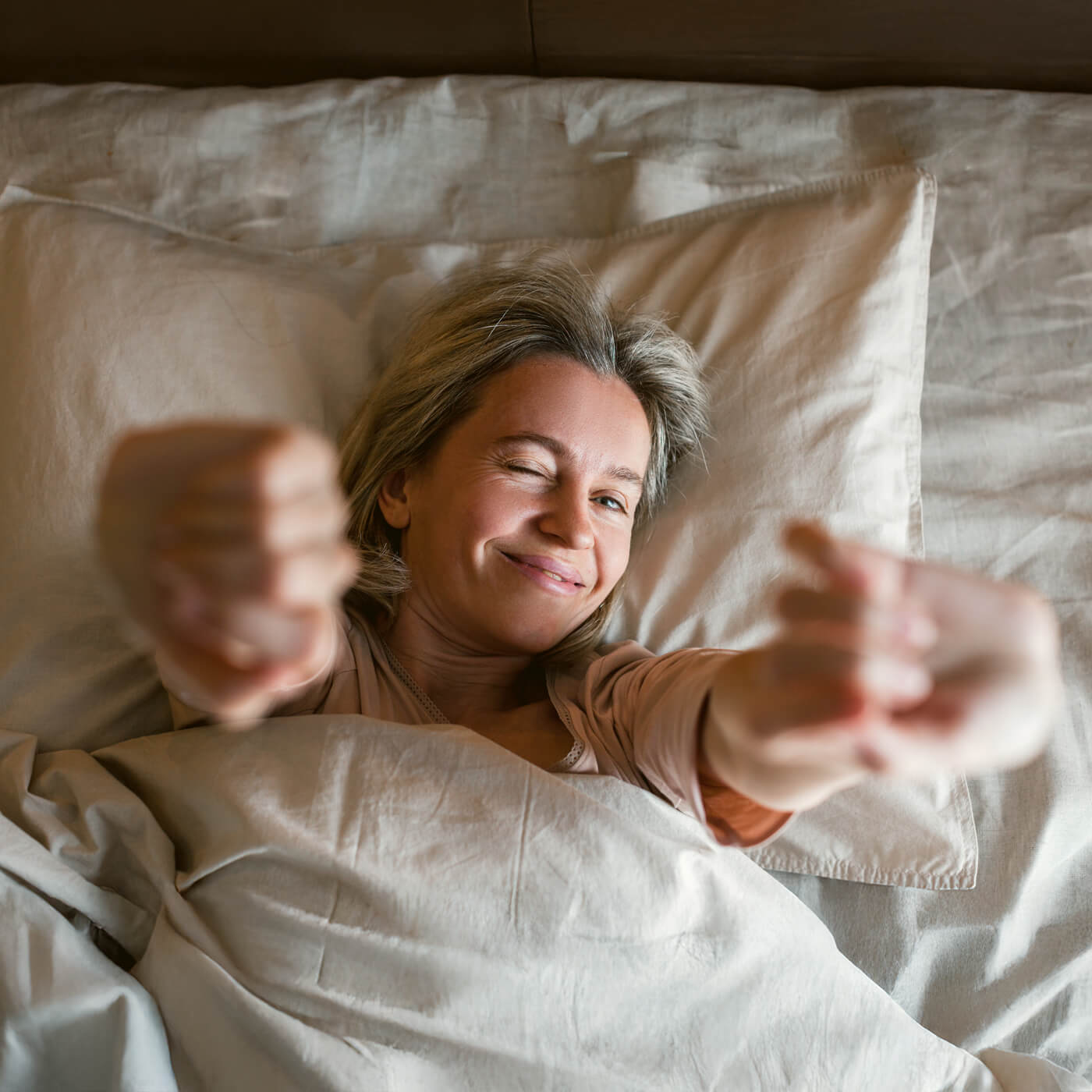 Middle-aged woman in bed smiling and stretching her arms while winking.