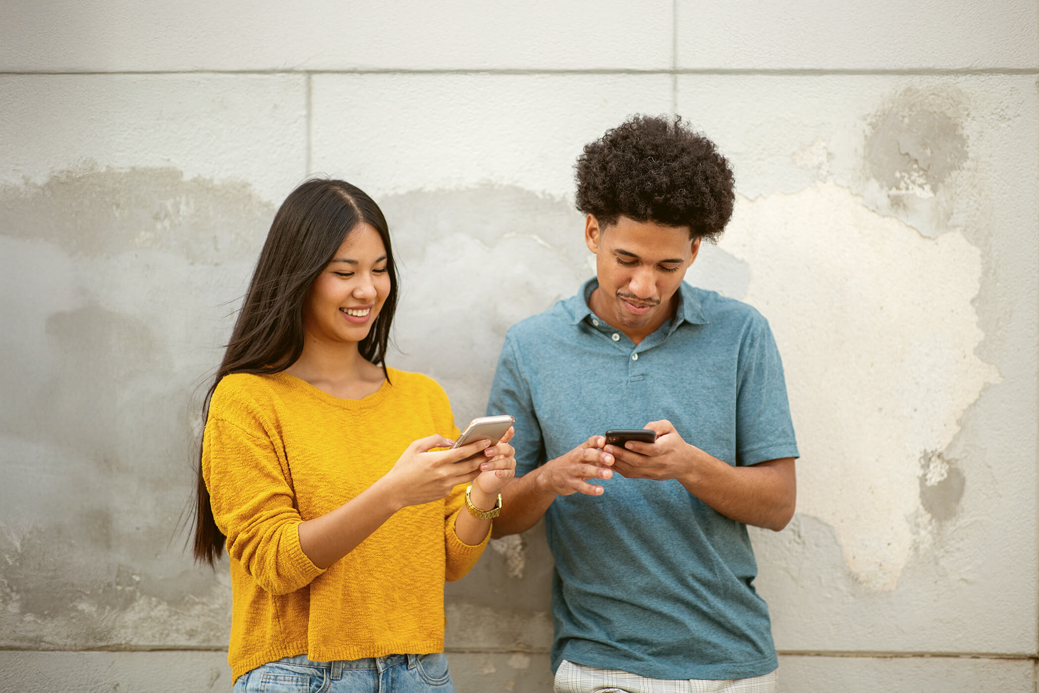 Smiling young woman in a yellow sweater and young man in a blue shirt standing against a worn wall, both looking at their smartphones.