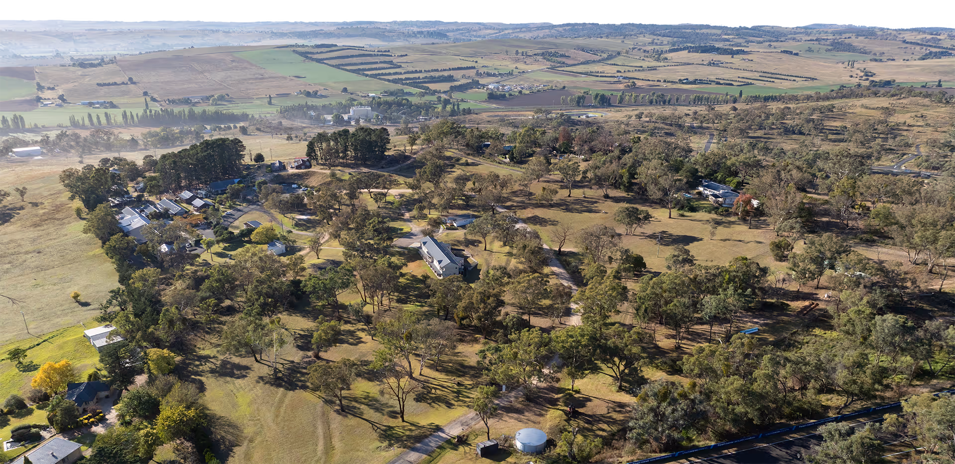 Aerial view of scattered rural houses surrounded by trees and open fields with farmland in the background.