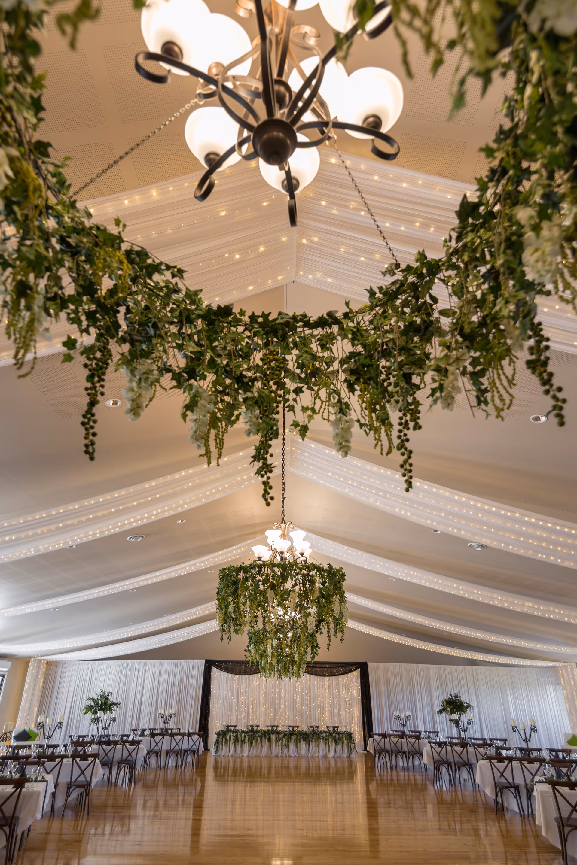 Elegantly decorated event hall with draped ceilings, chandeliers wrapped in greenery, and rows of tables with white tablecloths and chairs.