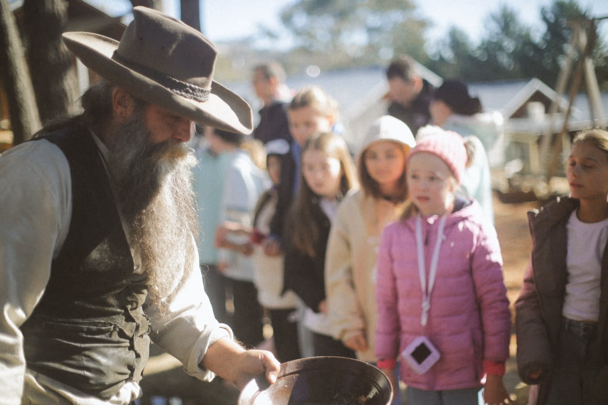 Bearded man in a wide-brimmed hat pan gold while children watch attentively in an outdoor setting.
