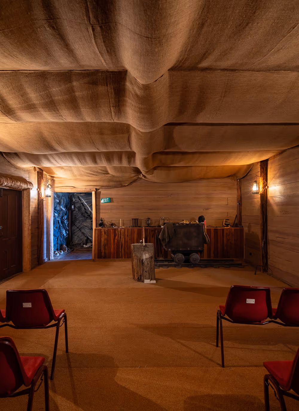 Indoor room with red chairs facing a wooden mining cart on rails and a candle on a wooden stump under a fabric-covered ceiling.
