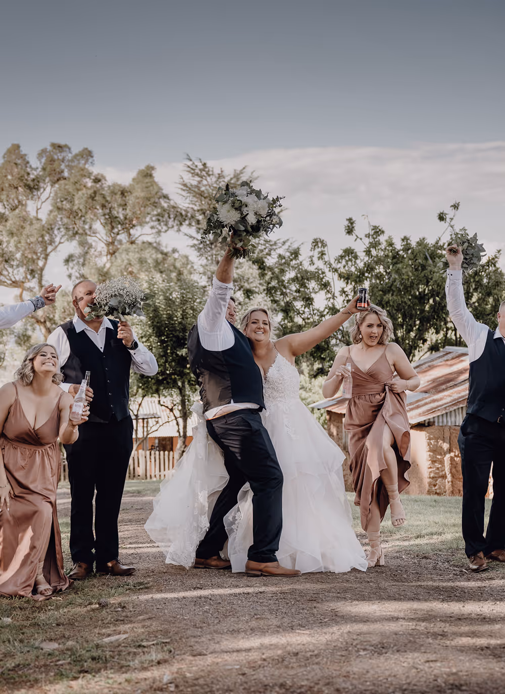 Bride and groom celebrating outdoors with bridesmaids and groomsmen, some holding bouquets and drinks.