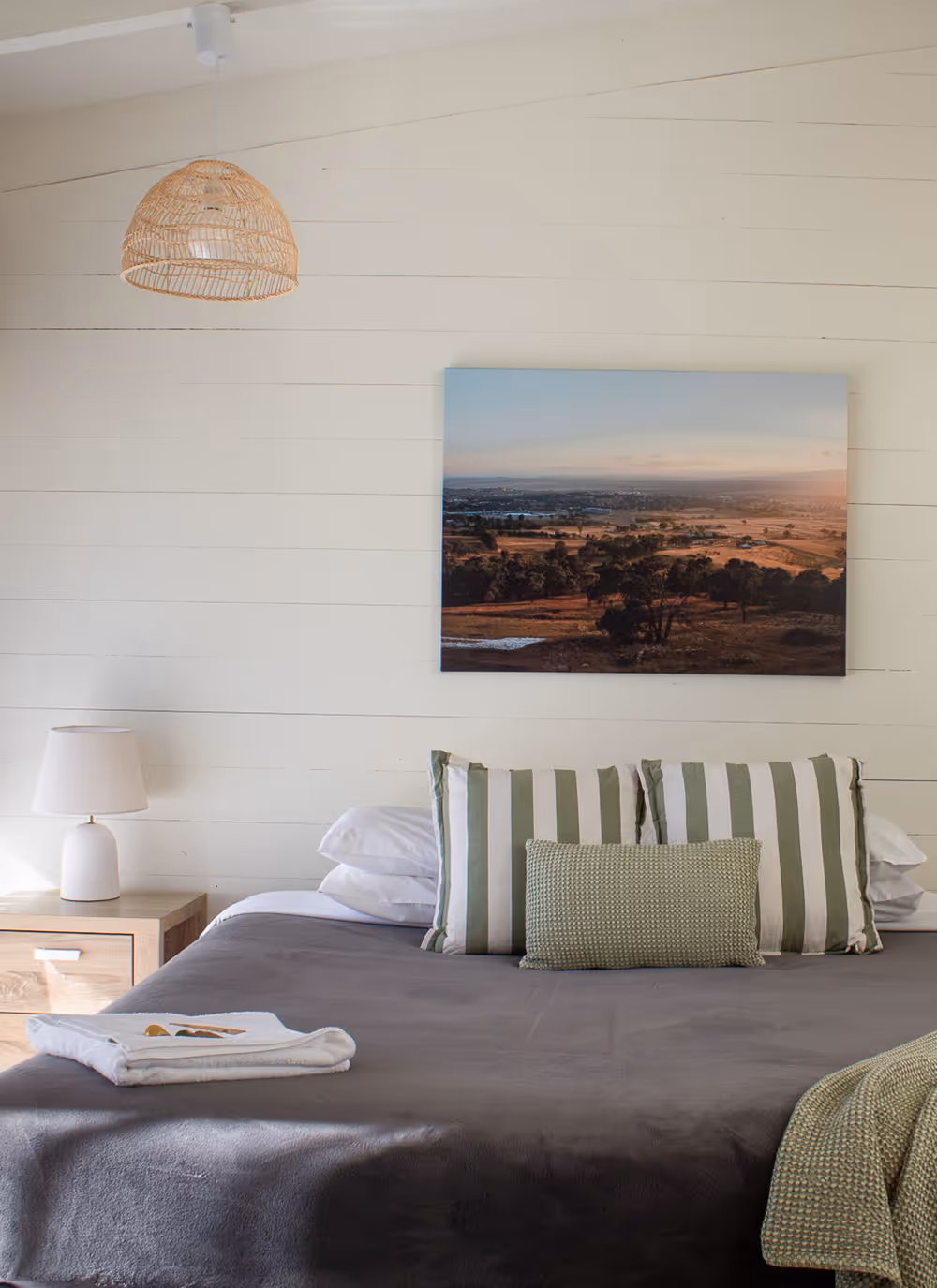 Minimalist bedroom with a gray bedspread, striped green and white pillows, a bedside lamp on a wooden nightstand, and a landscape photo on the wall.